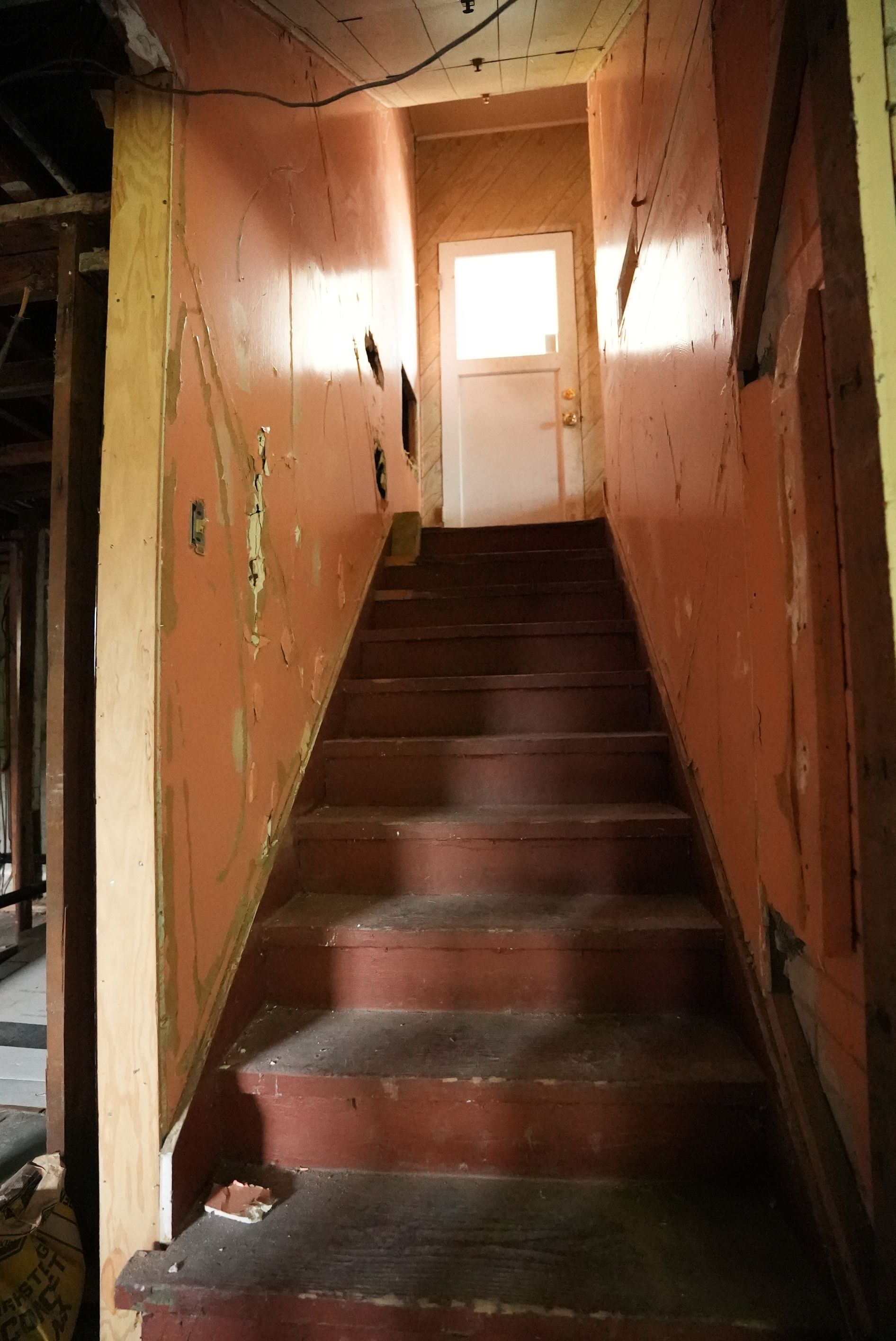 Narrow dim stairway with worn dark steps and peeling peach walls leading to a white door at the top