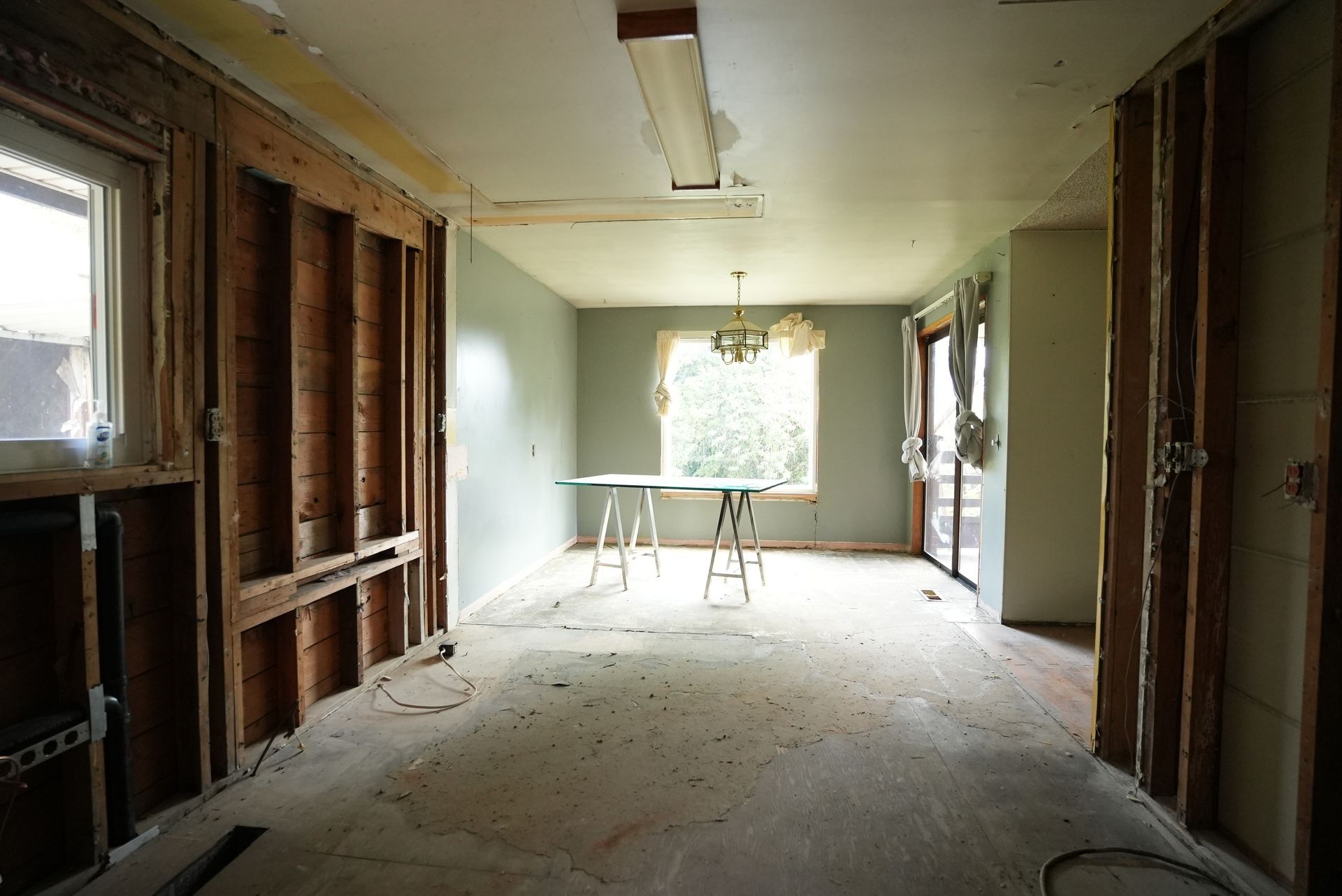 Interior of a stripped-down room under renovation, with exposed studs, debris, and sunlight through a window.