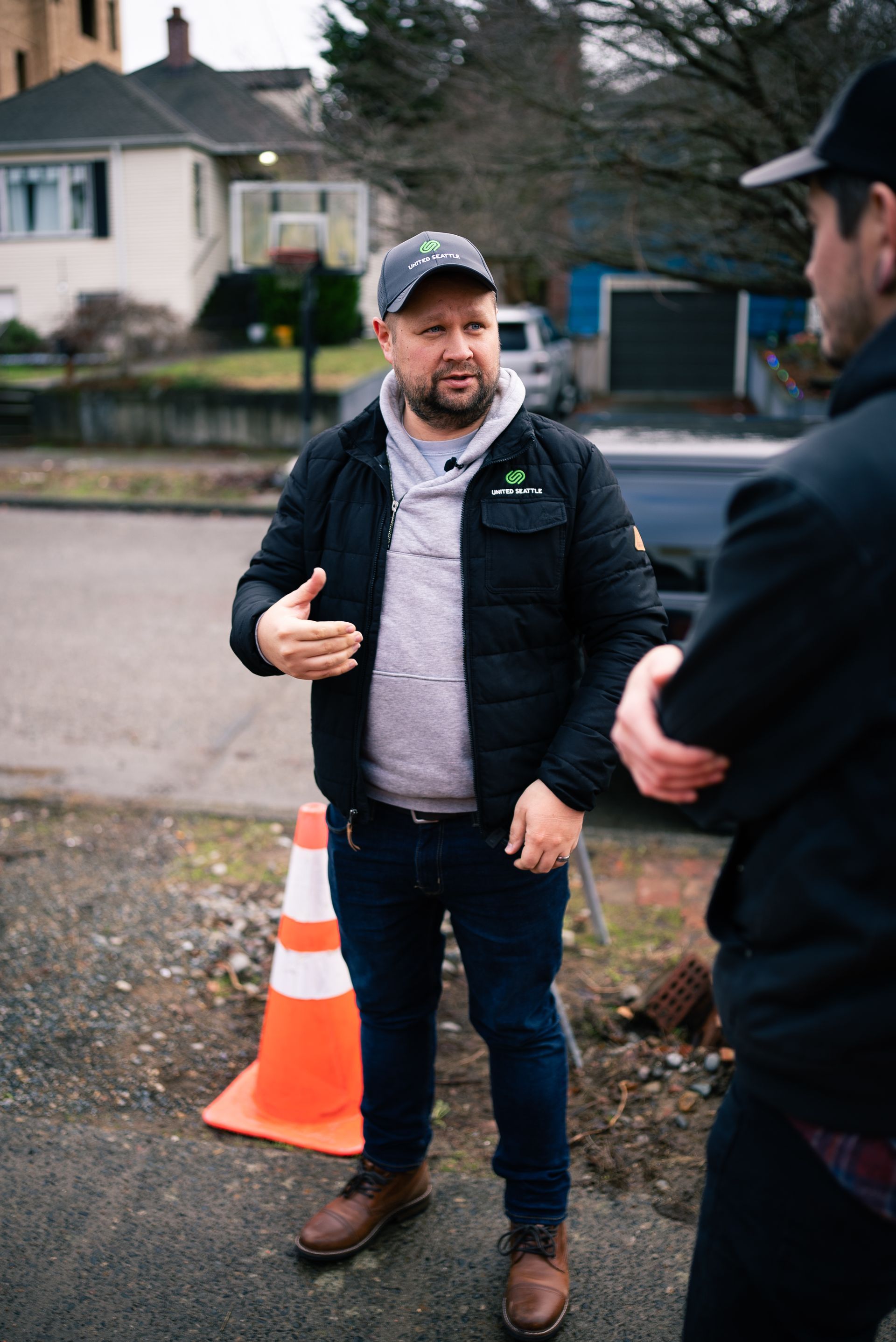 Man in a cap speaking outdoors near a traffic cone, with another person in the foreground.