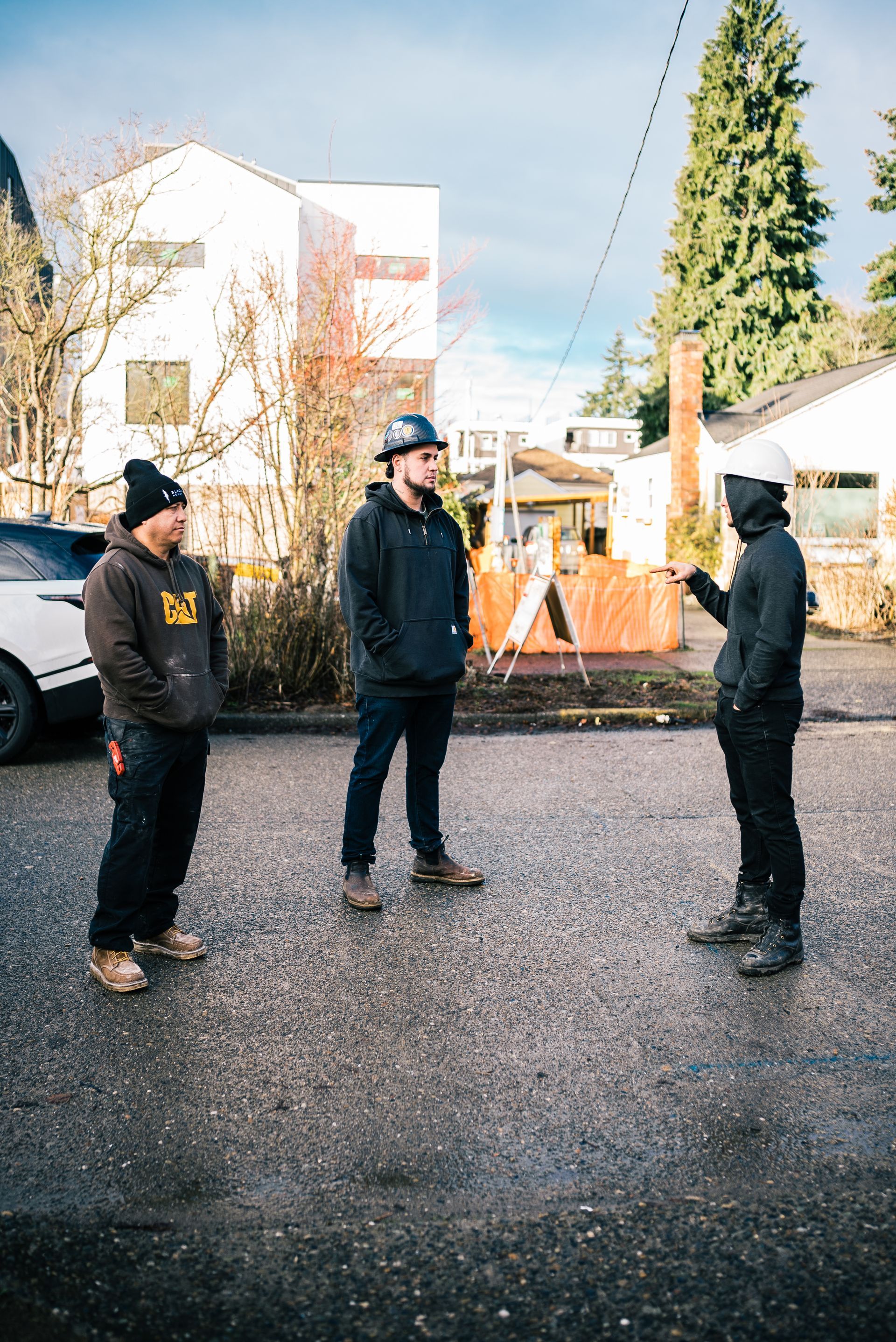 Three people stand talking in a gravel driveway beside houses and trees.