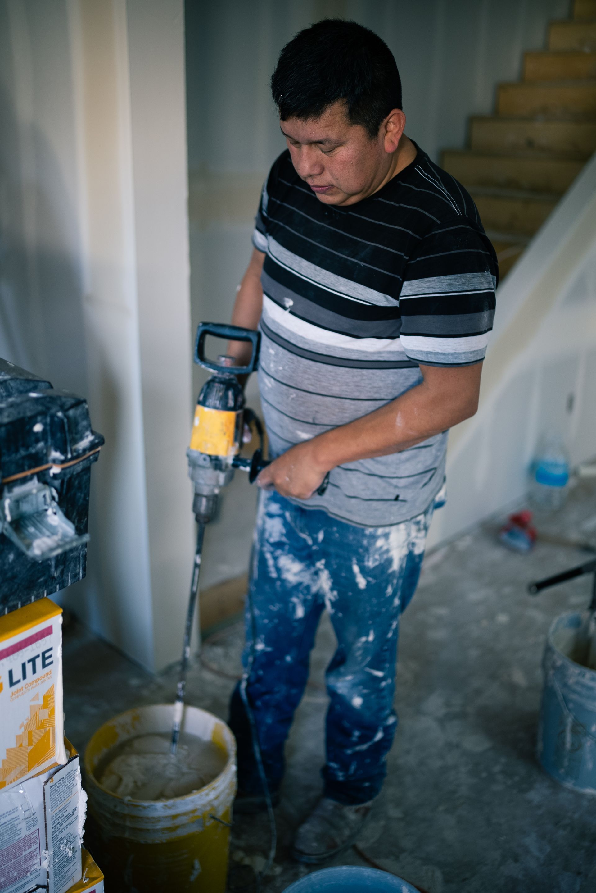 Man in paint-splattered clothes using a drill in a room under renovation