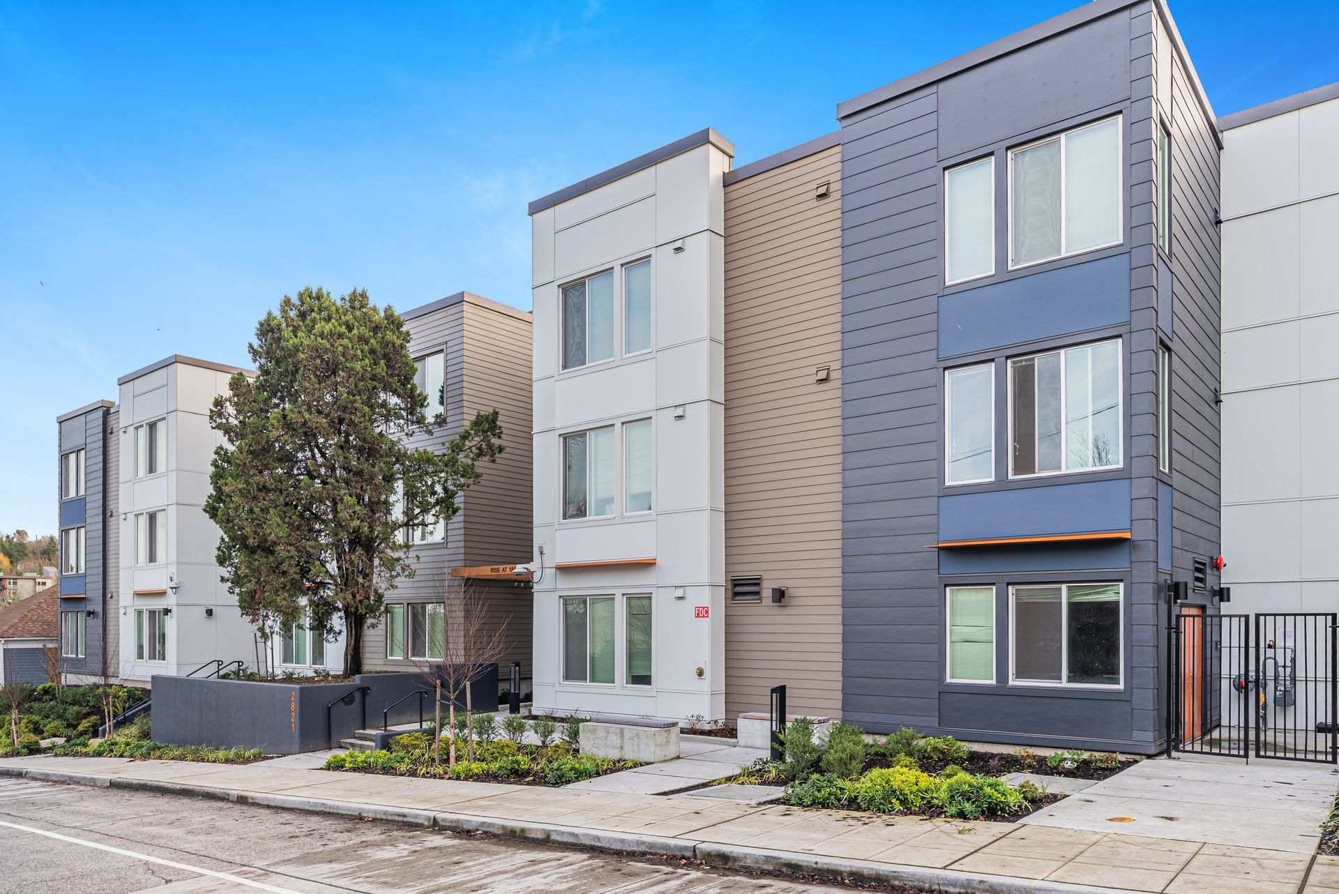 Row of modern apartment townhouses with gray facades, balconies, and a tree-lined sidewalk under a blue sky