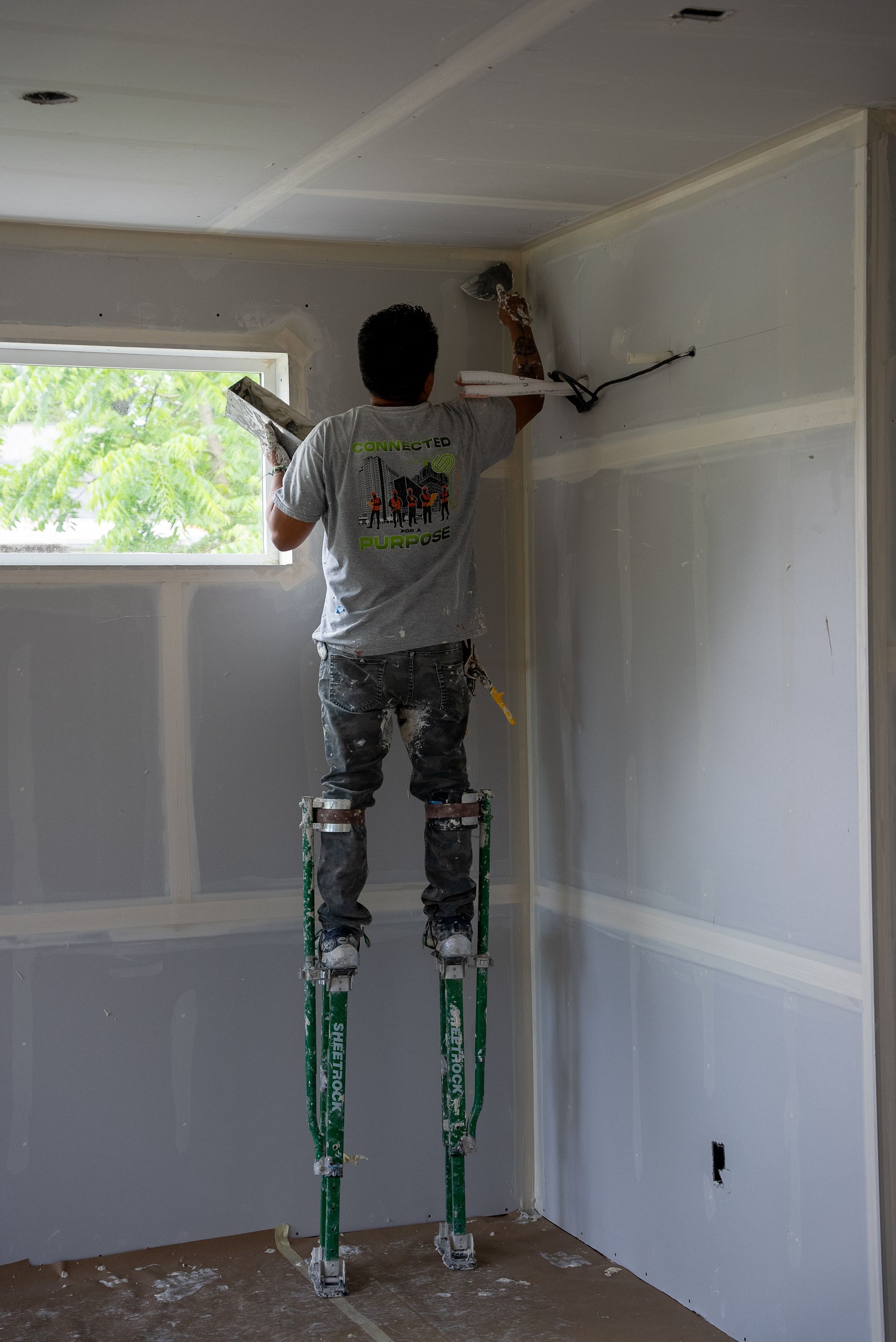 Person painting a room’s upper wall and ceiling with a roller on long extension poles