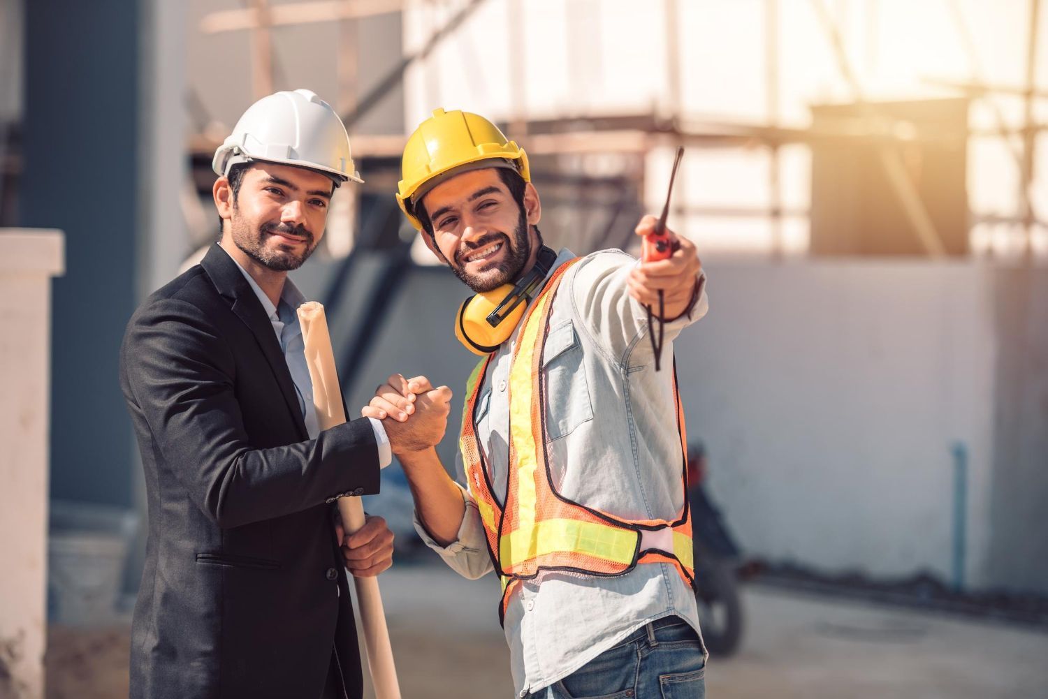 Two construction workers in hard hats standing together at a job site, one pointing ahead and smiling.