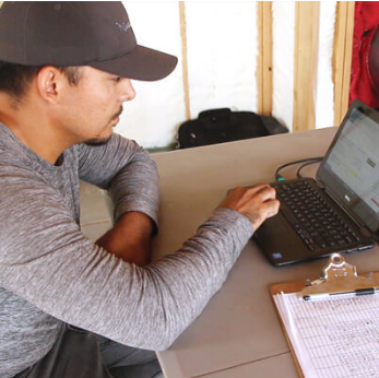 Person at a table pointing at a laptop beside a clipboard and notebook.