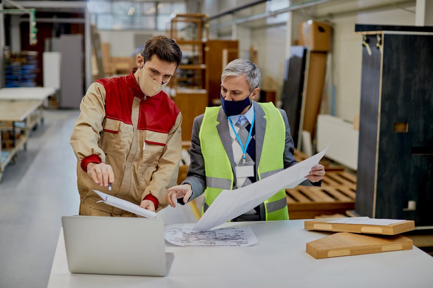 Two men wearing face masks are looking at a blueprint in a factory.