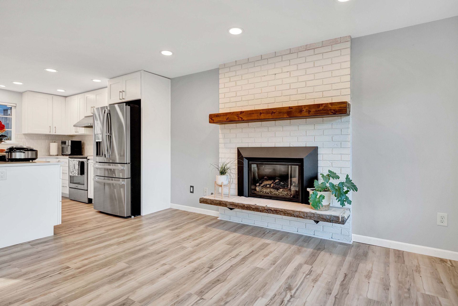Open-concept living room with white brick fireplace, wood mantel, gray walls, and adjacent kitchen