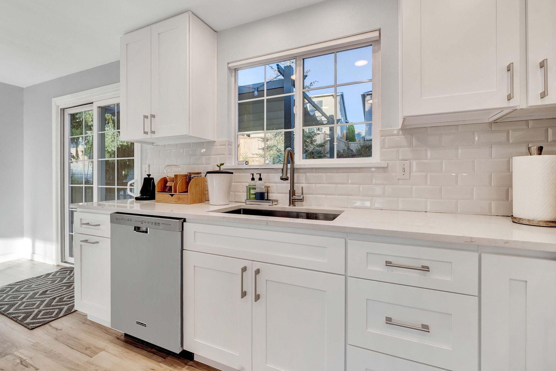 Bright white kitchen with sink, window, dishwasher, and white cabinets against a subway tile backsplash