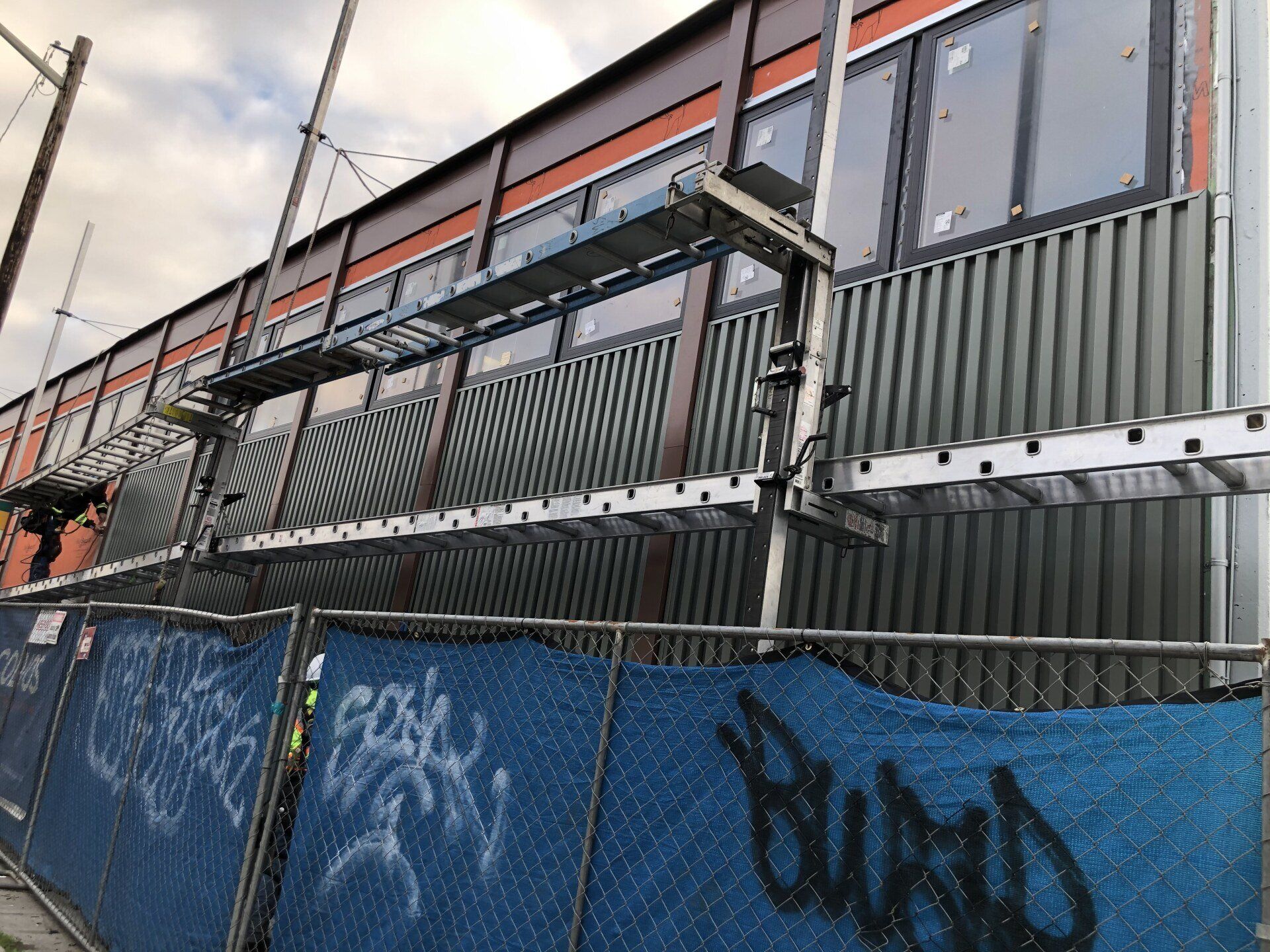 Blue construction fence in front of a corrugated metal building with scaffolding and graffiti