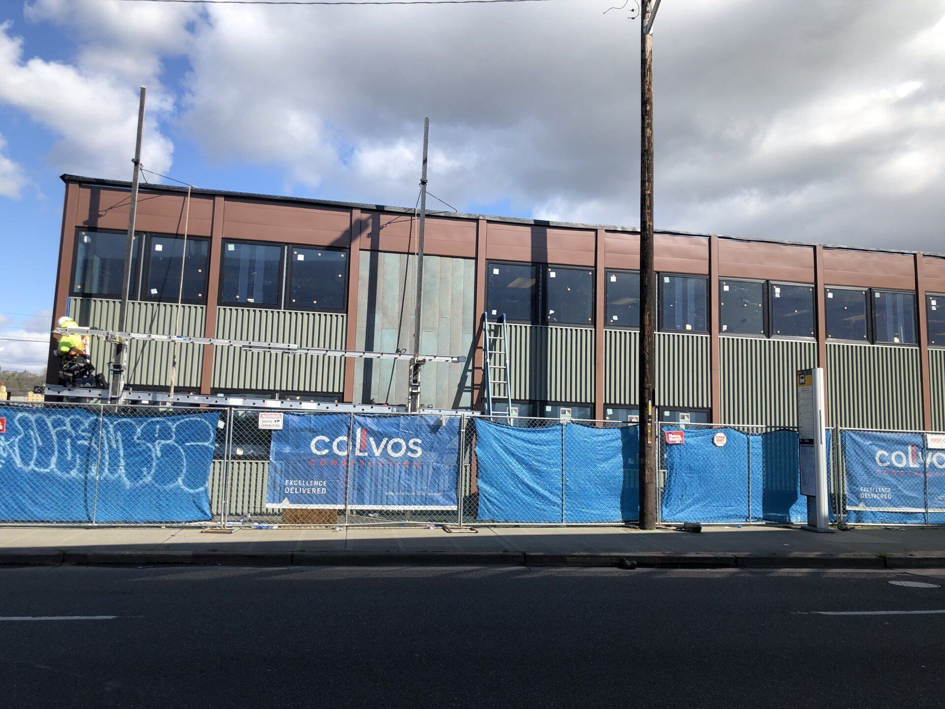 Blue construction fencing in front of a partially built brick-and-glass building under a cloudy sky