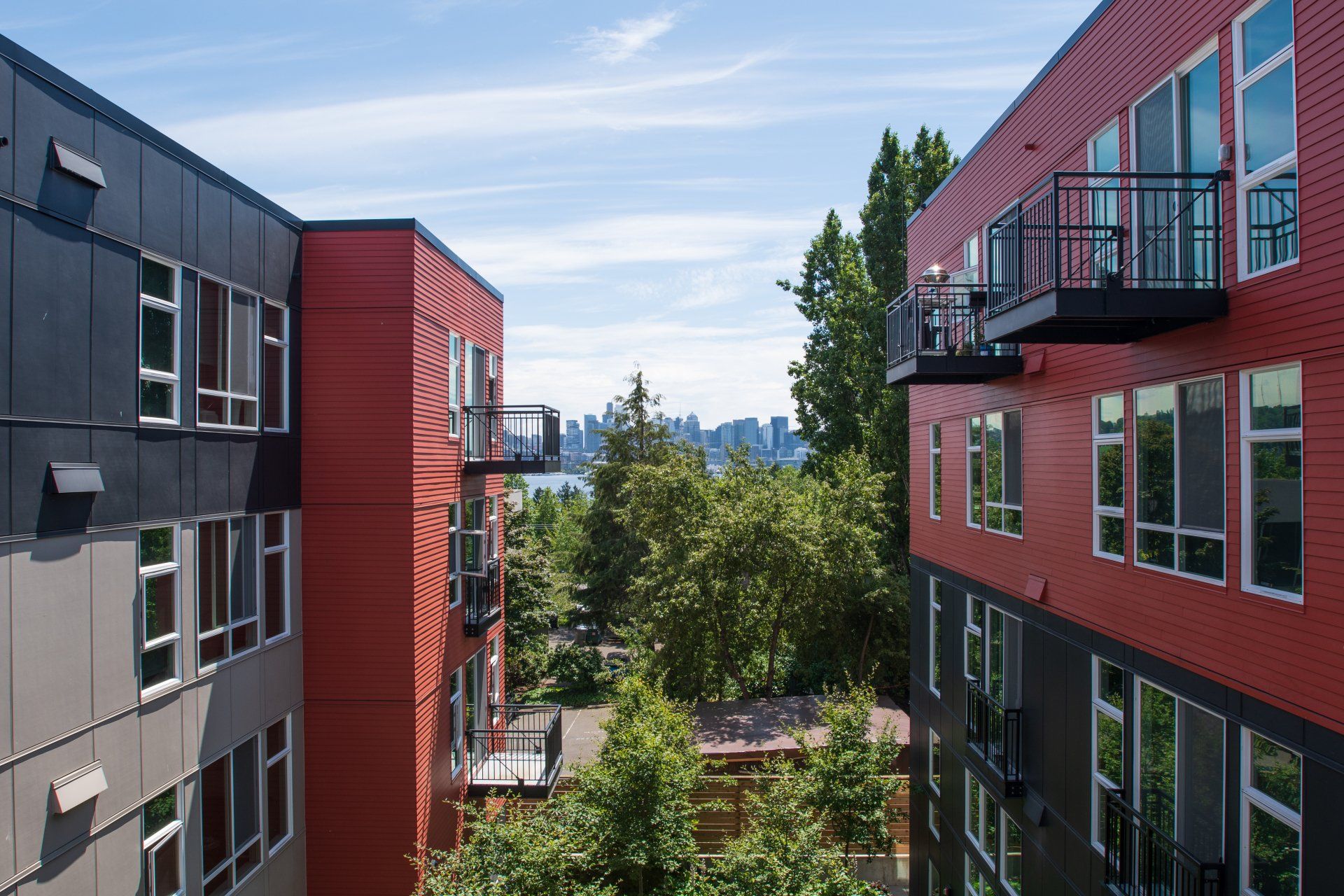 Modern apartment buildings with balconies framing a green courtyard and distant city view under a blue sky