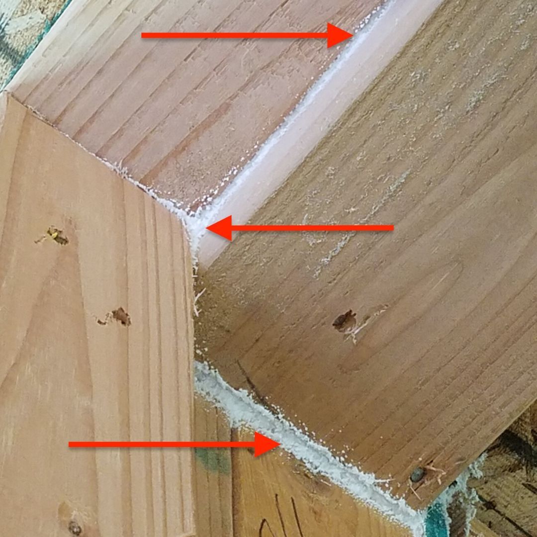 Close-up of a wooden corner joint with white adhesive or filler highlighted by red arrows.