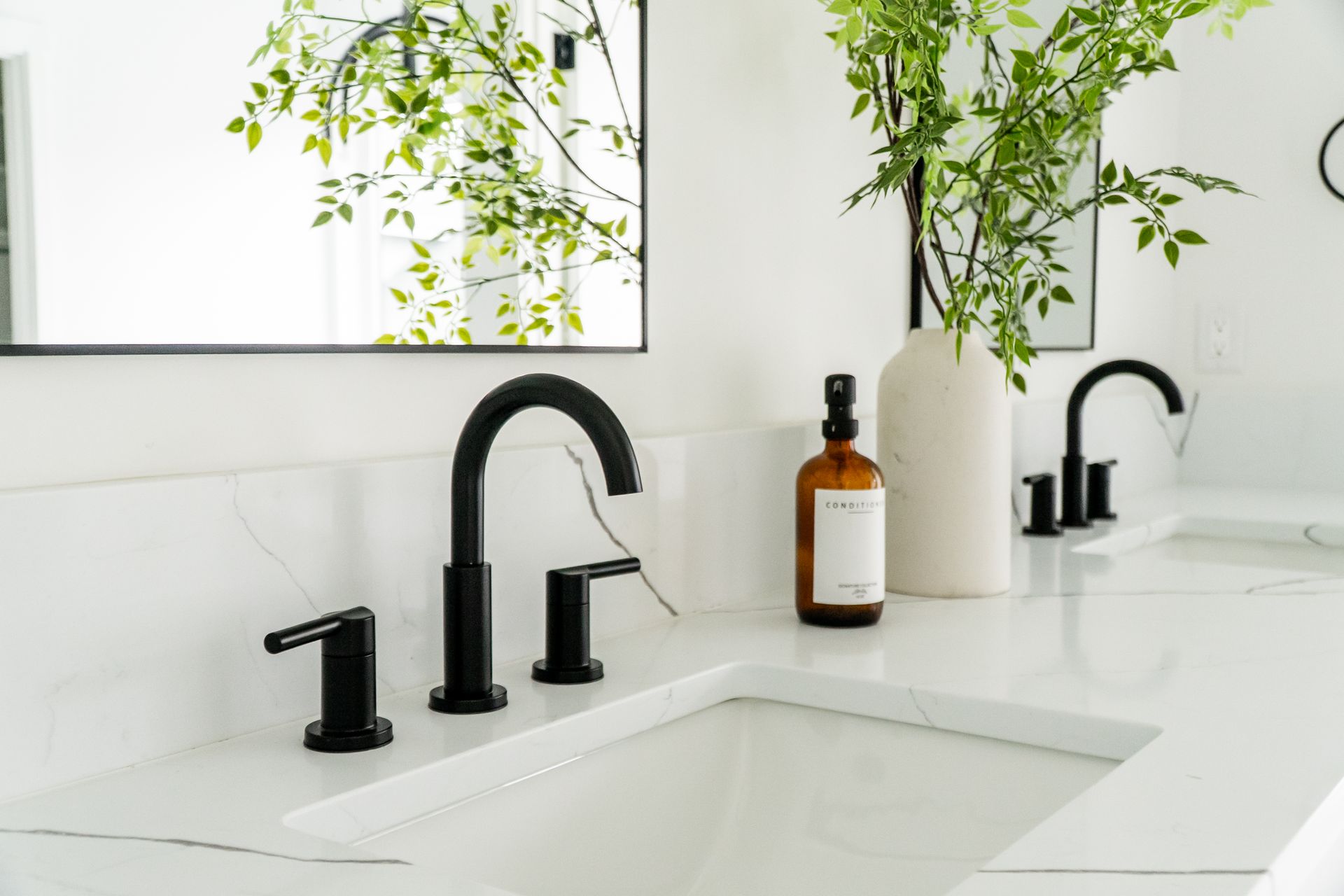 Modern white bathroom sink with black faucet, soap dispenser, mirror, and green plants.