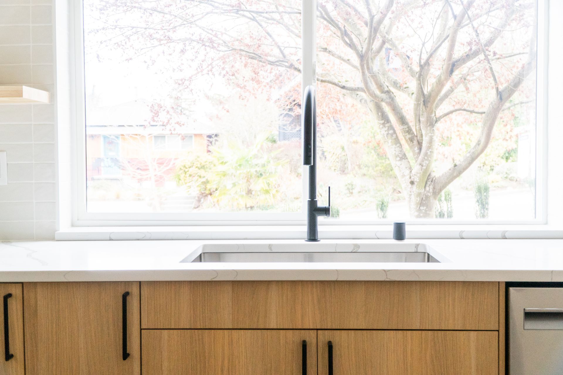 Modern kitchen sink beneath large window, with wood cabinets and a black faucet.