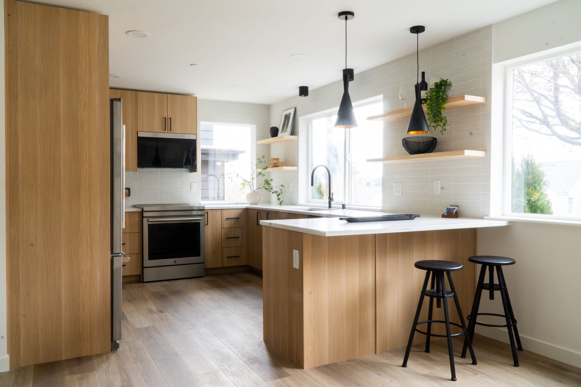 Bright modern kitchen with wood cabinets, white island, pendant lights, and two stools by a window.