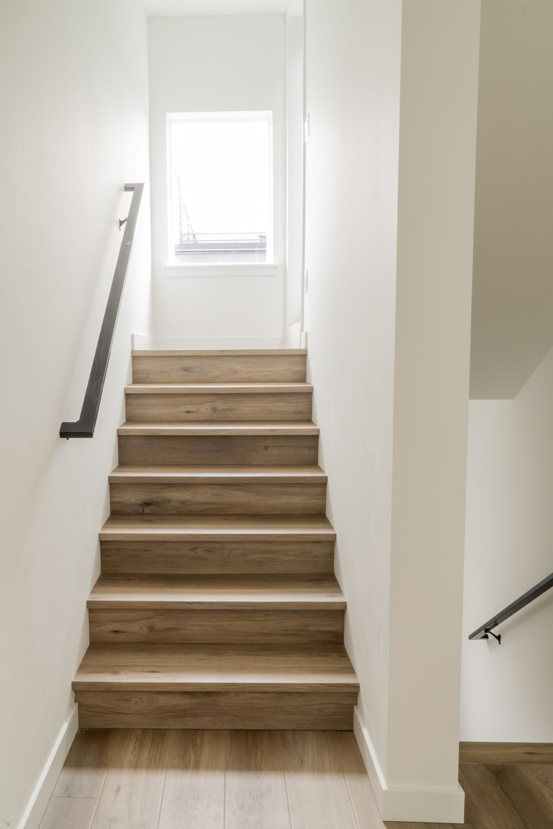 Staircase with wood steps and black handrails leading to a bright window in a white hallway