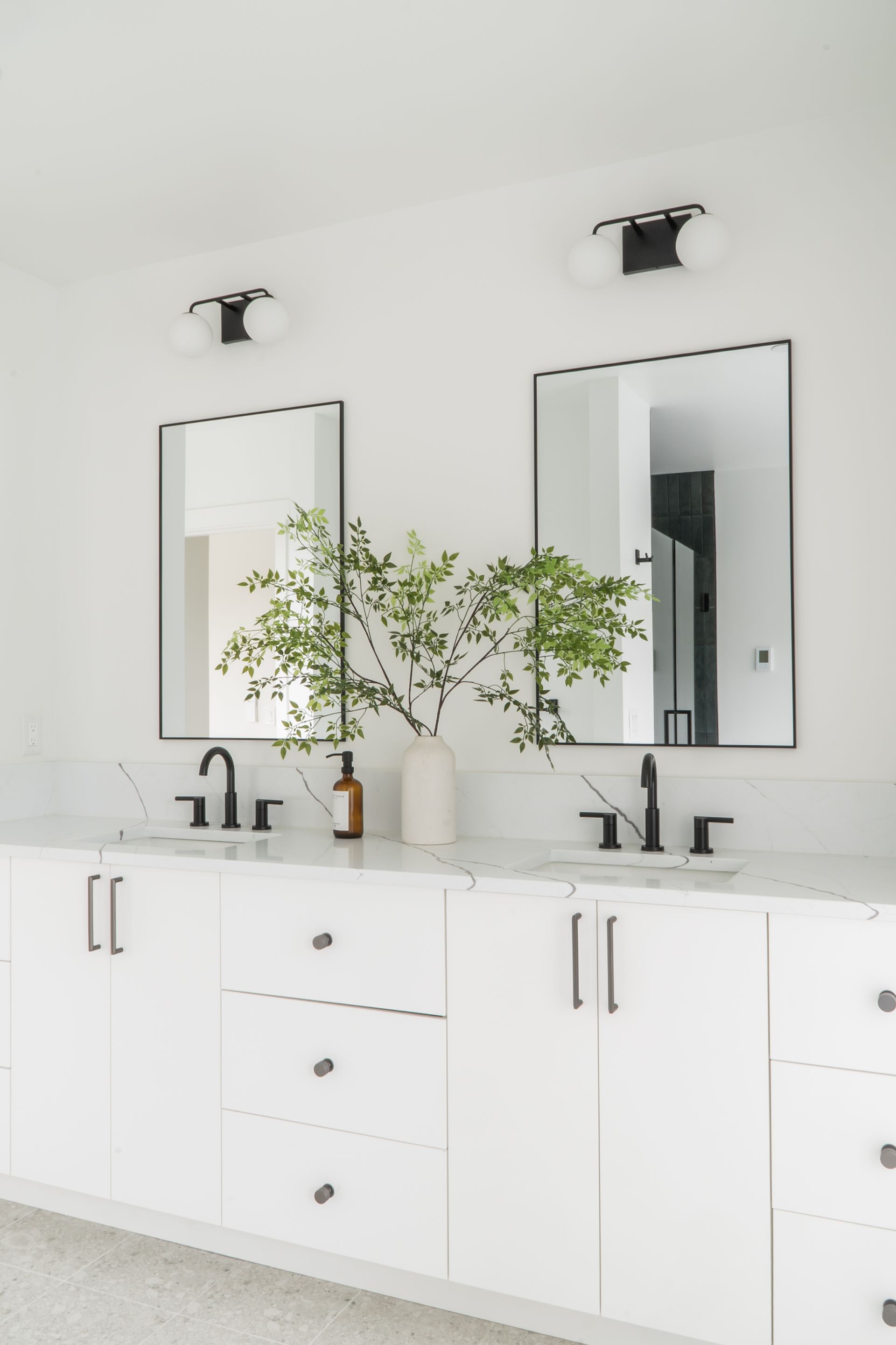 Minimalist white bathroom vanity with black fixtures and two mirrors, with a plant in the center