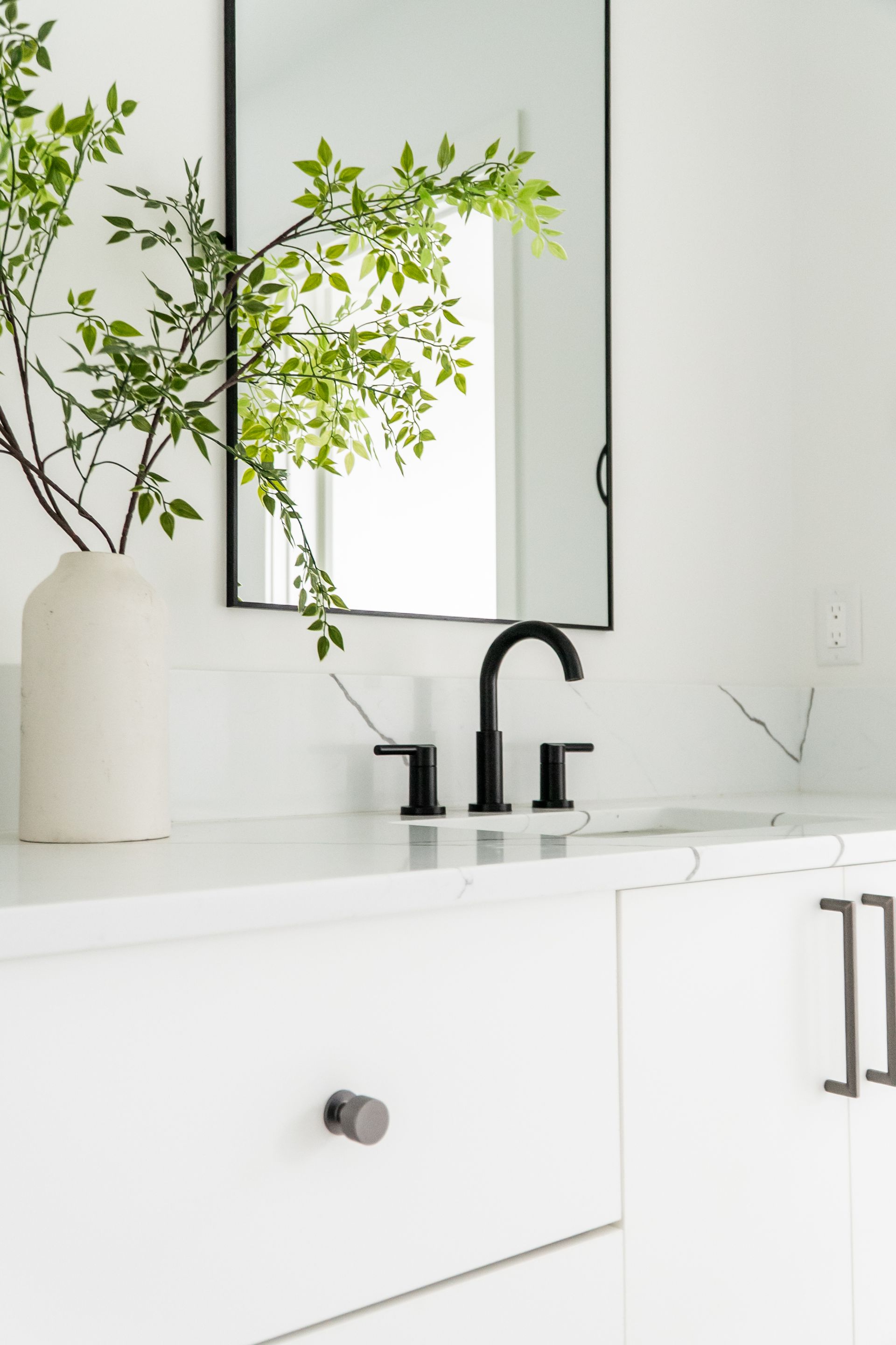 Minimal white bathroom vanity with black faucet, mirror, and green plant in a vase