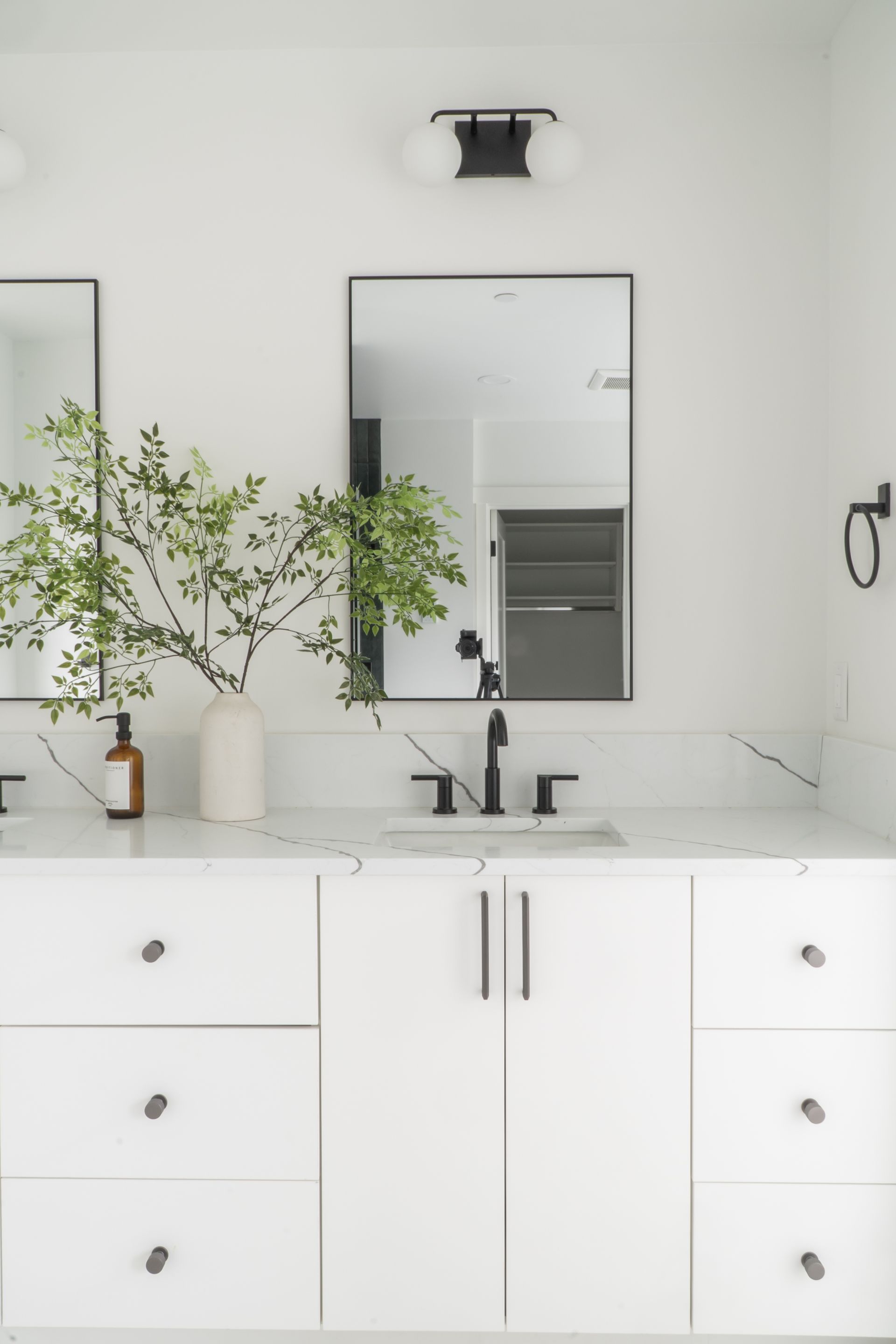 Modern white bathroom vanity with double sinks, mirror, wall light, and potted greenery