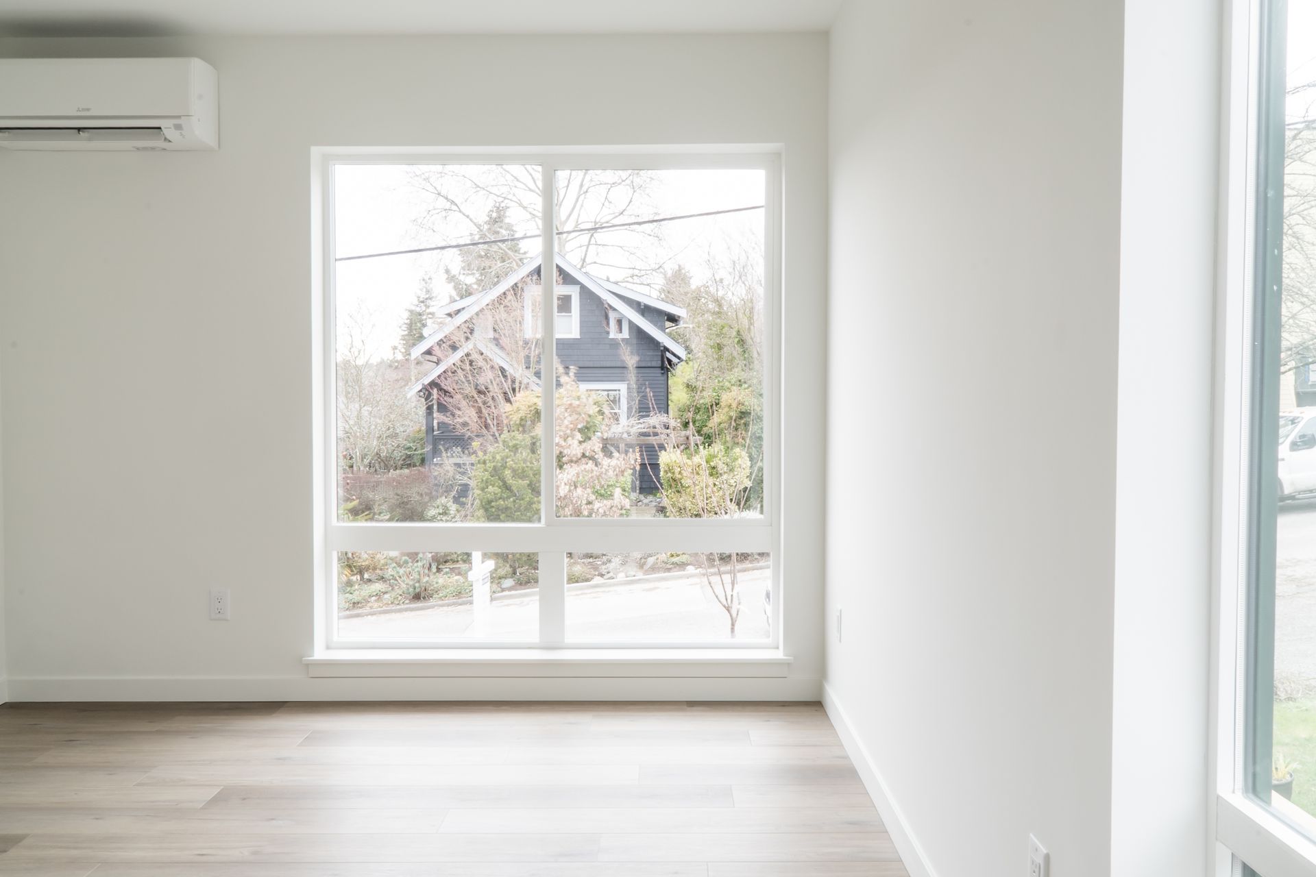 Bright empty room with large window, white walls, light wood floor, and an air conditioner.
