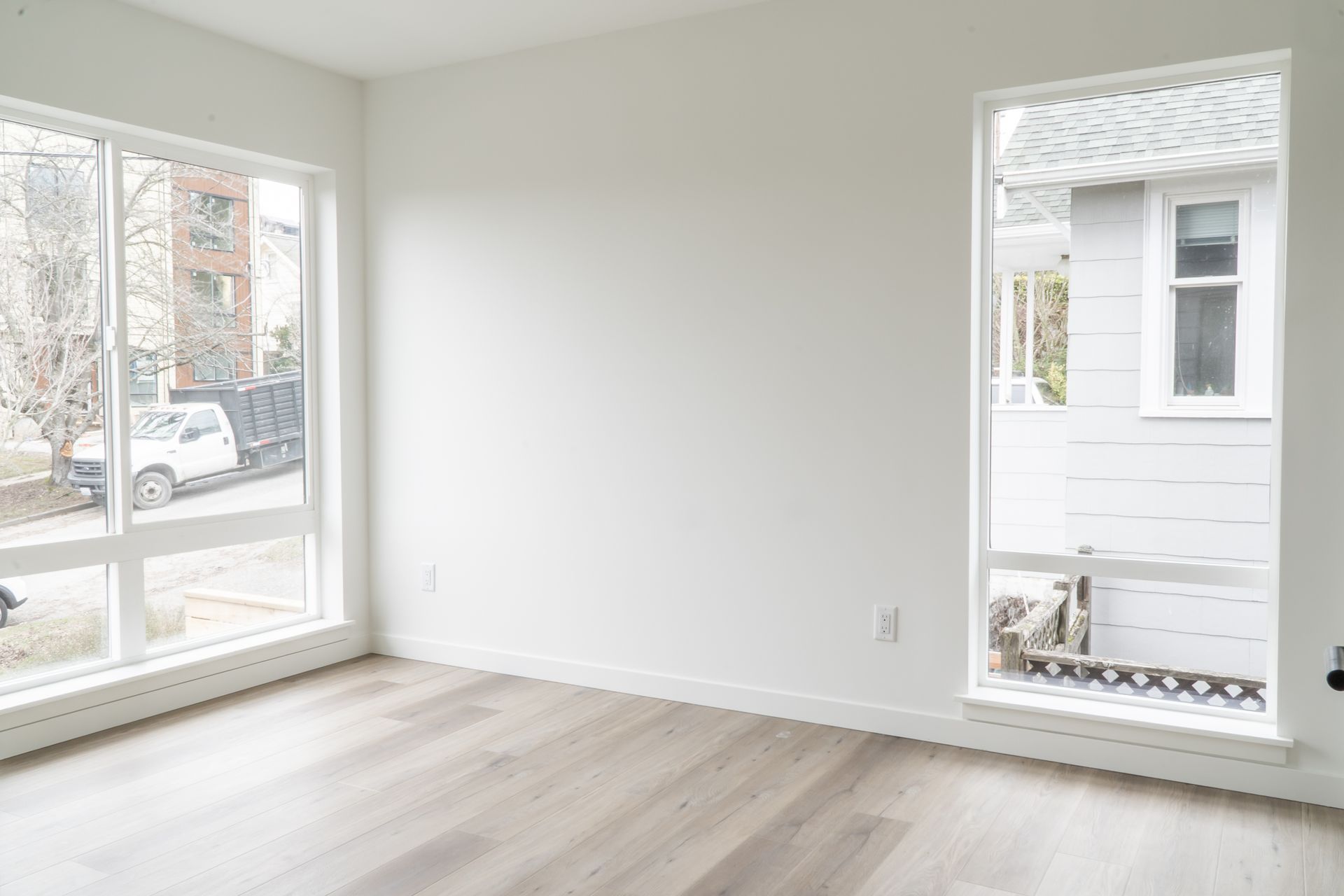 Empty bright room with white walls, light wood floor, and large windows overlooking neighboring buildings