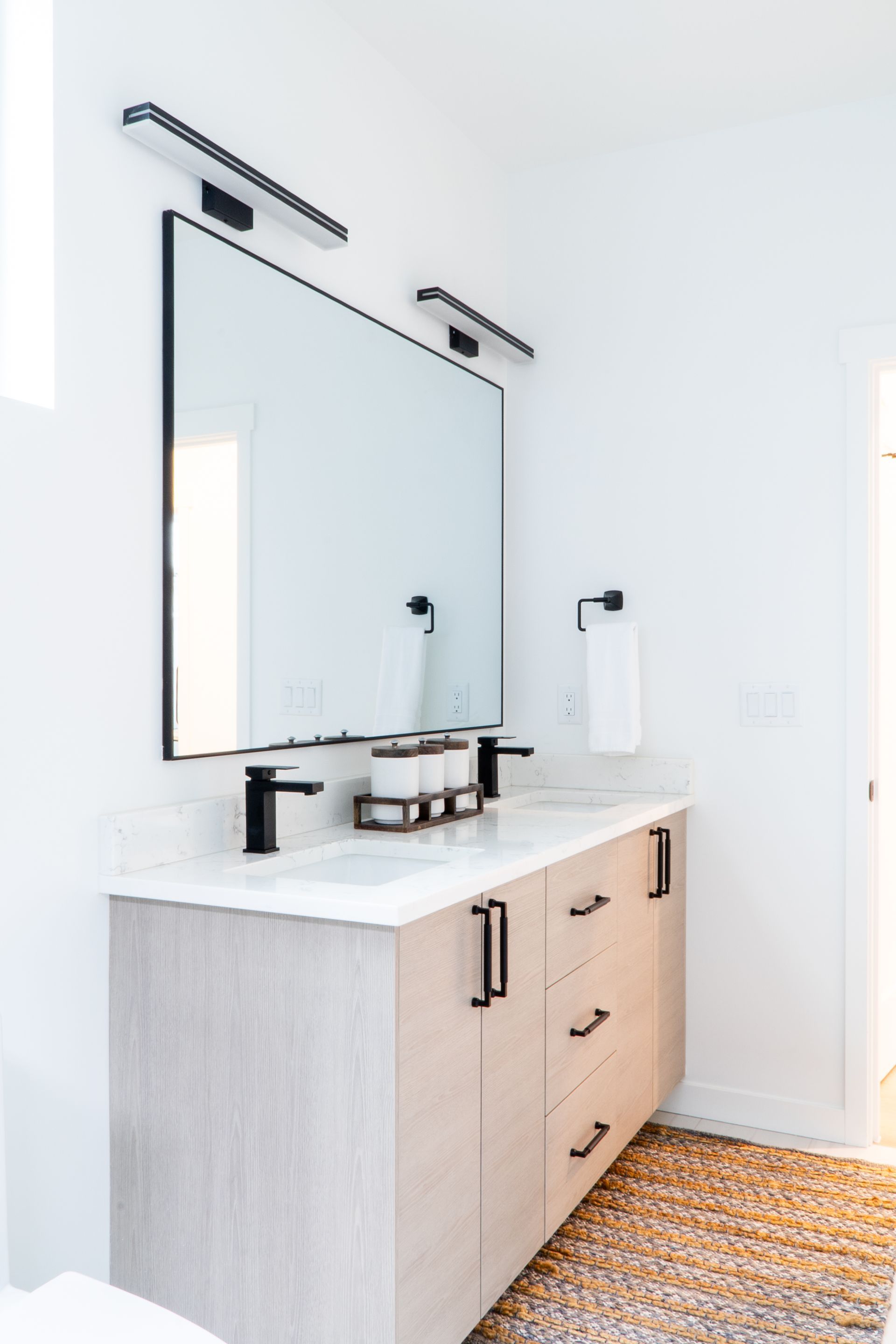 Bright modern bathroom vanity with large mirror, black fixtures, and light wood cabinet on patterned rug.