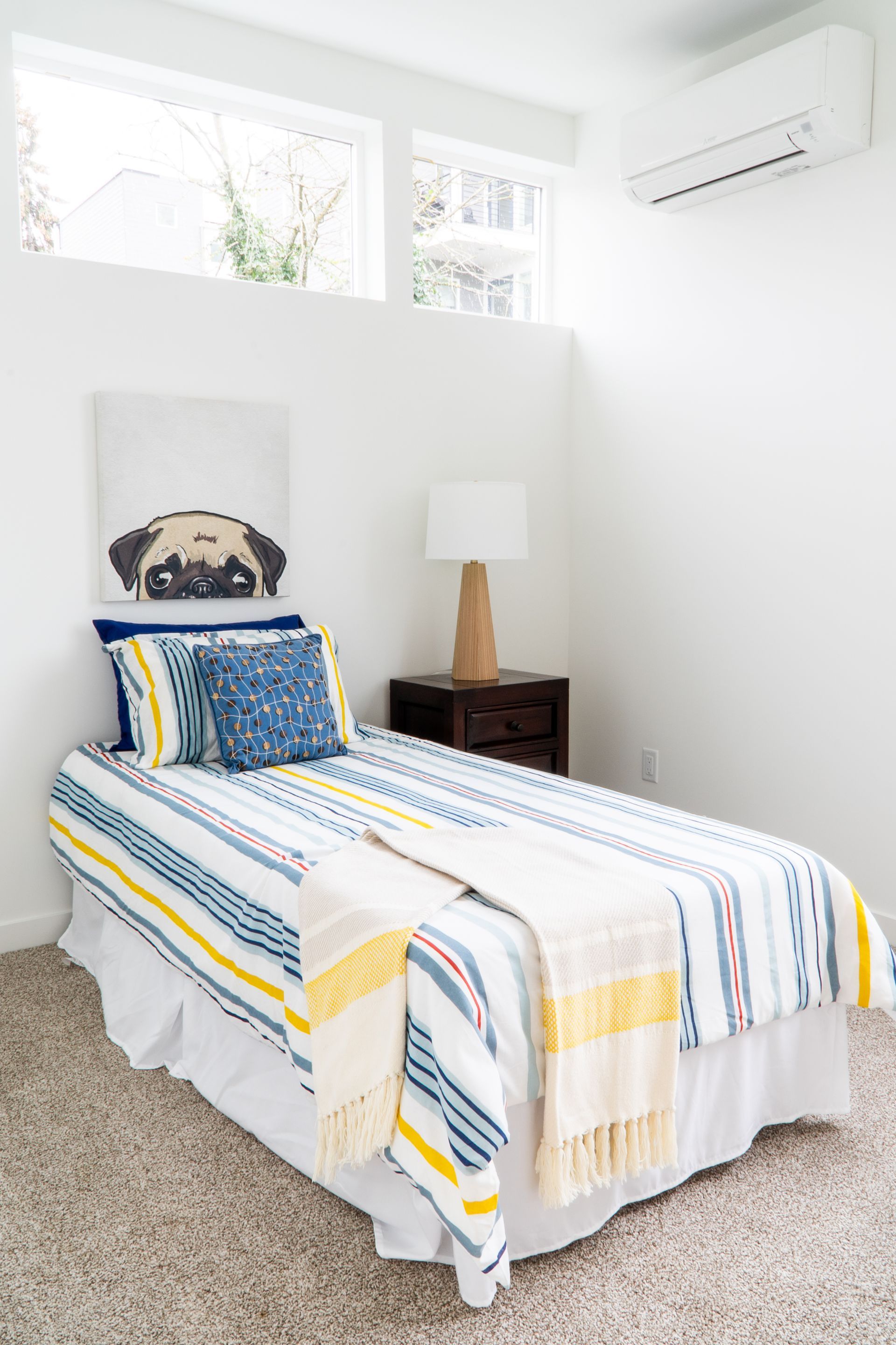 A small bedroom with a neatly made twin bed, striped bedding, bedside lamp, and high windows.
