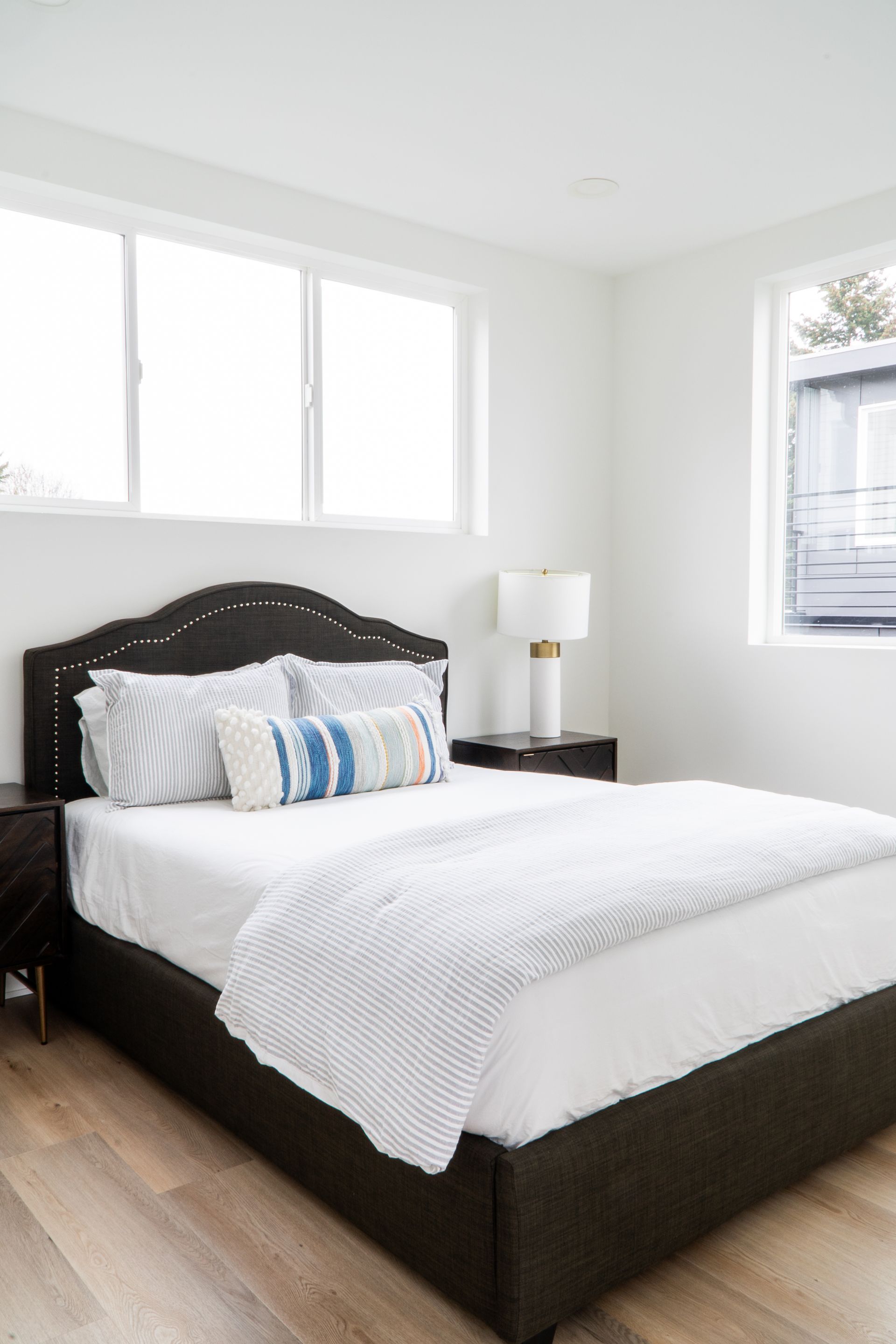 Bright bedroom with a dark bed frame, white bedding, and a bedside lamp near large windows.