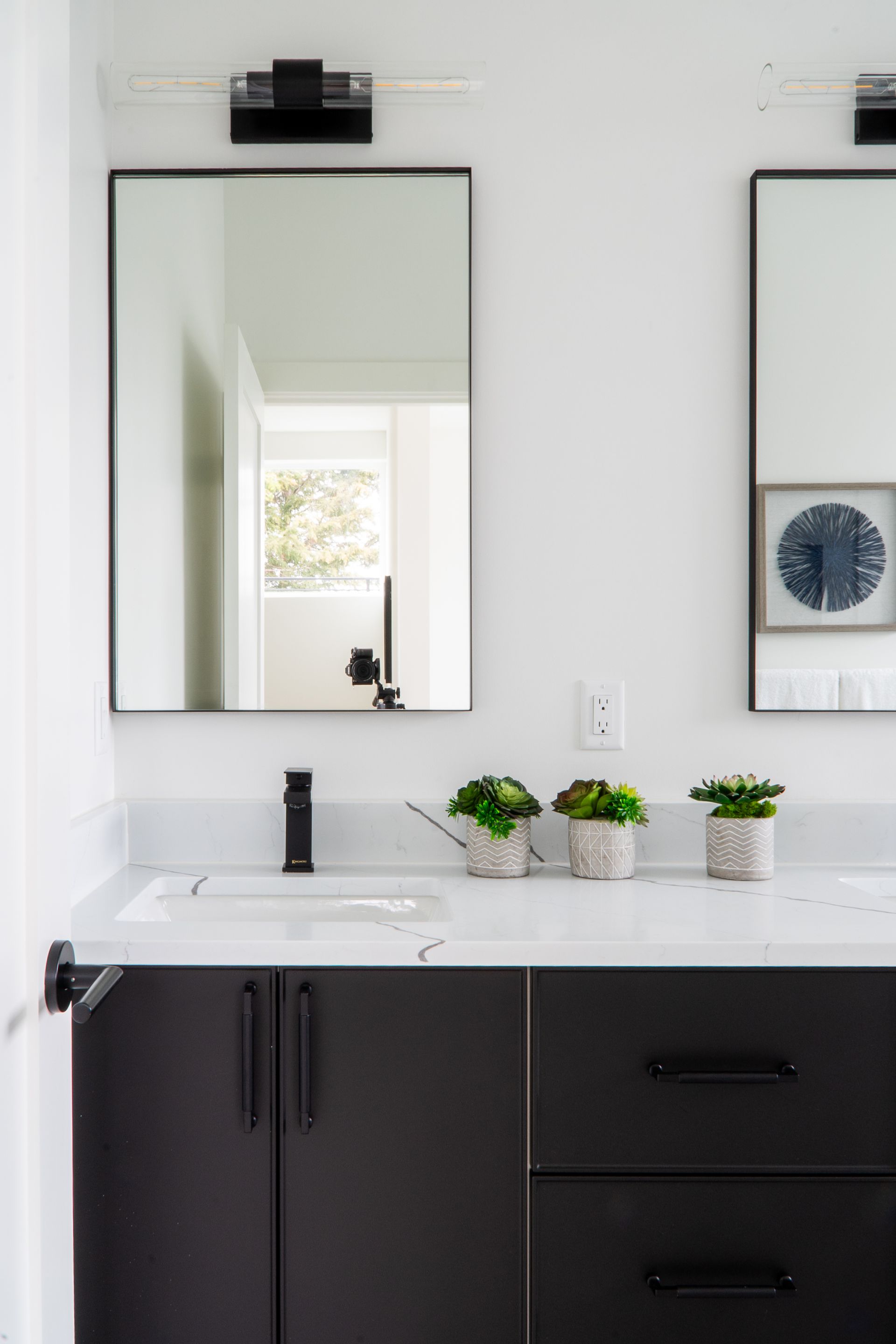 Modern bathroom vanity with black cabinets, mirror, wall light, and small potted plants on a white countertop