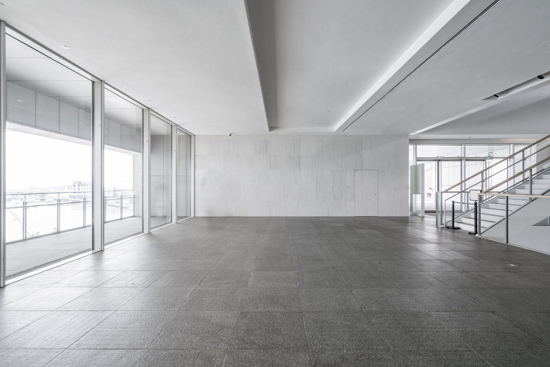 Modern empty interior with concrete floor, large windows, staircase, and white walls