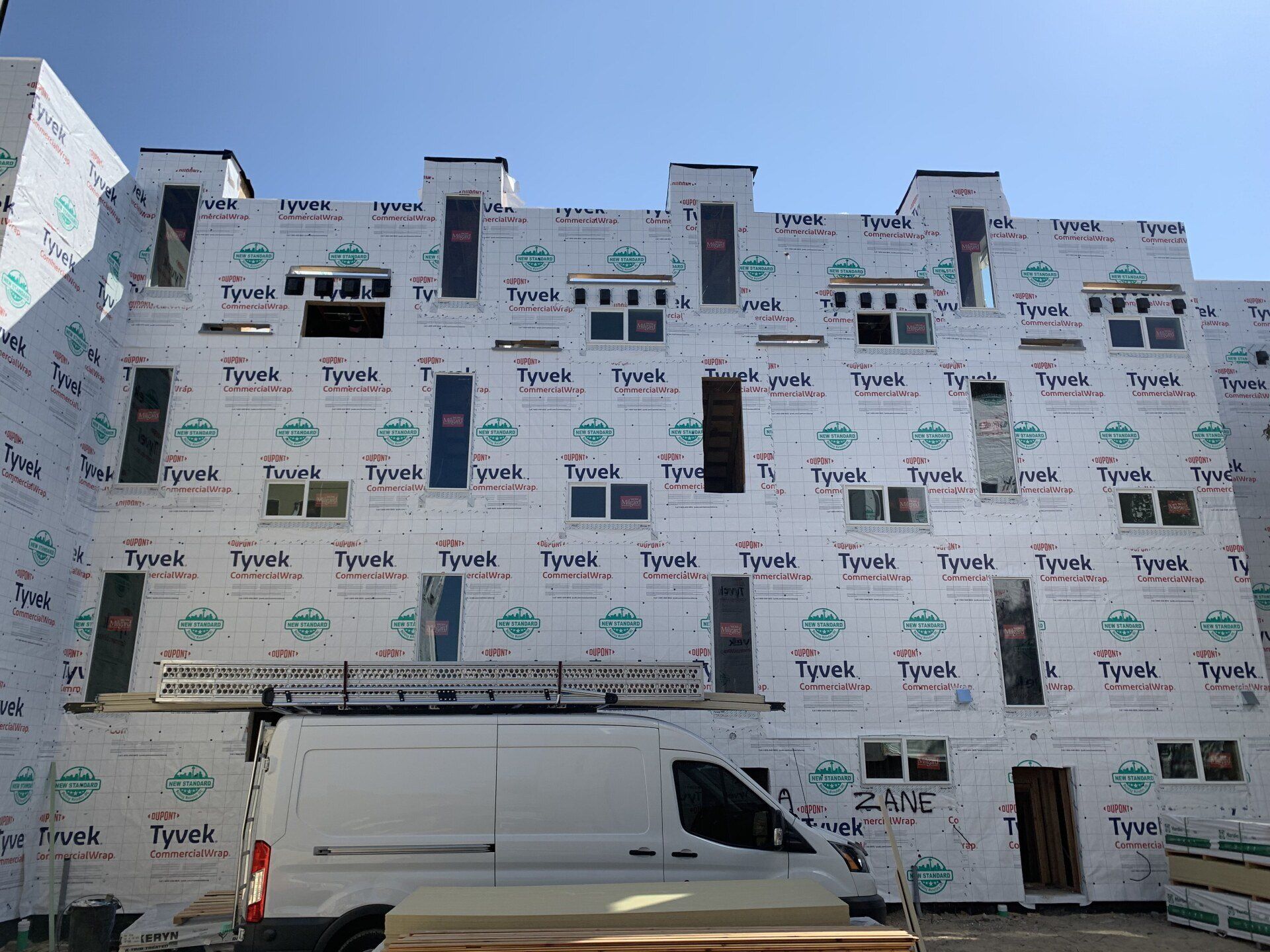 Partially covered building wrapped in white Tyvek house wrap, with a white van parked in front.