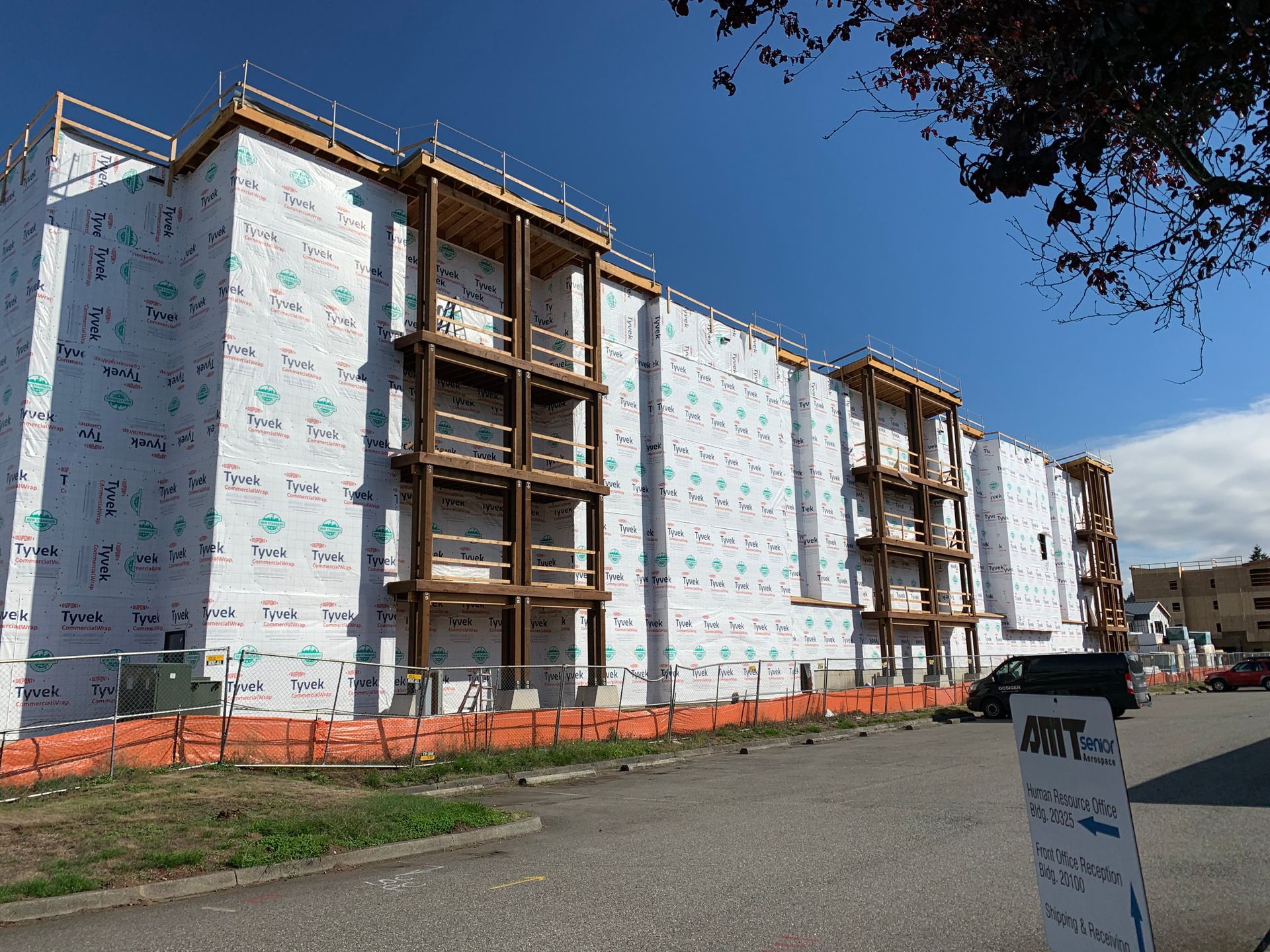 Row of apartment buildings under construction, wrapped in white sheathing with orange safety fencing and blue sky.
