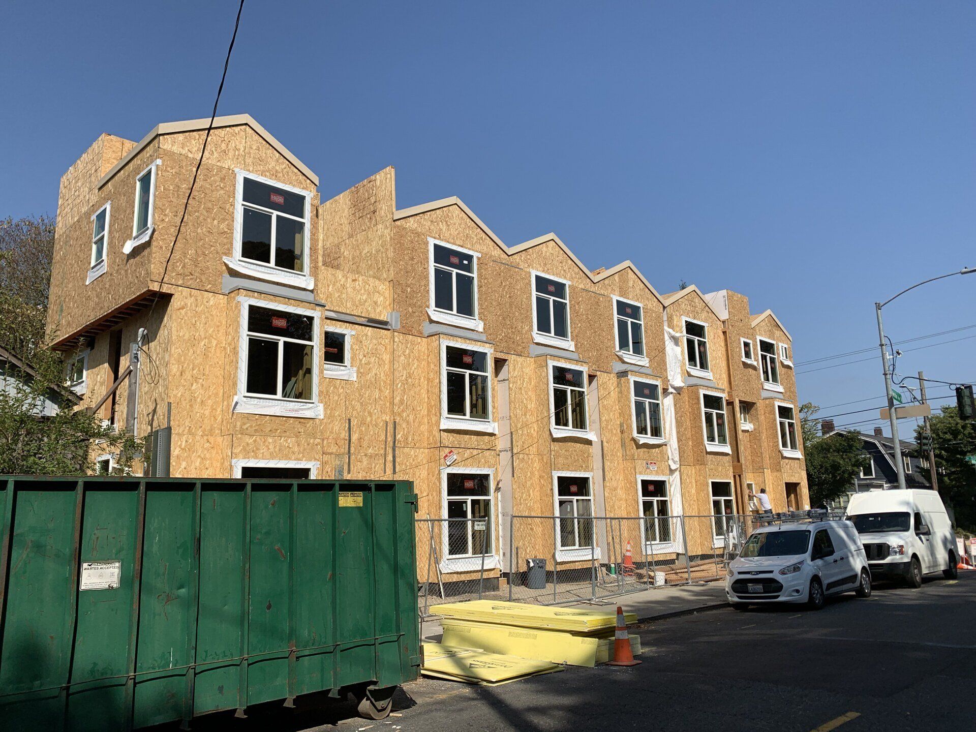 Partially built brick apartment building with green fence and parked vans on a street under a blue sky