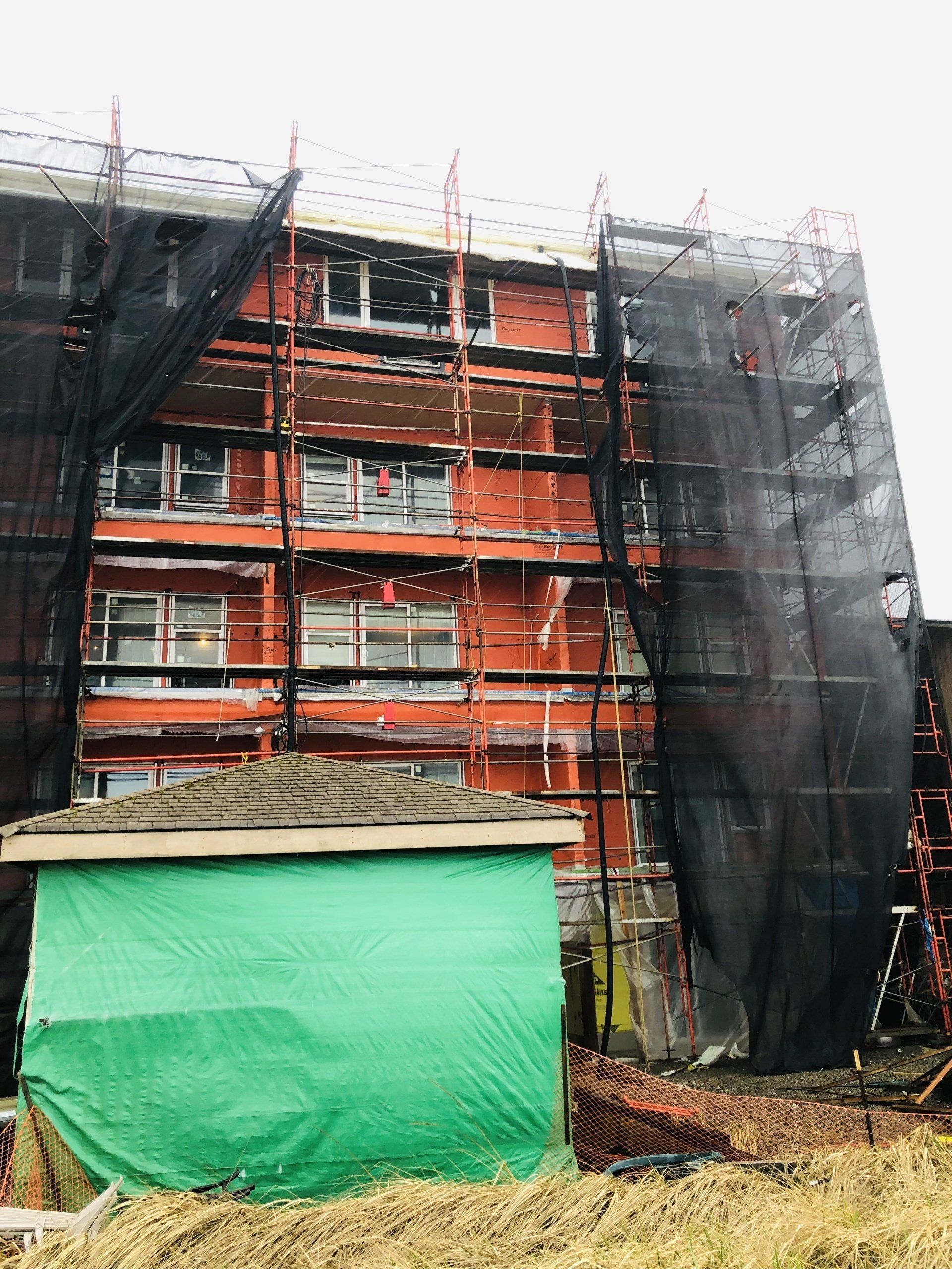 Multi-story building under construction, wrapped in black safety netting with a green-covered structure in front.