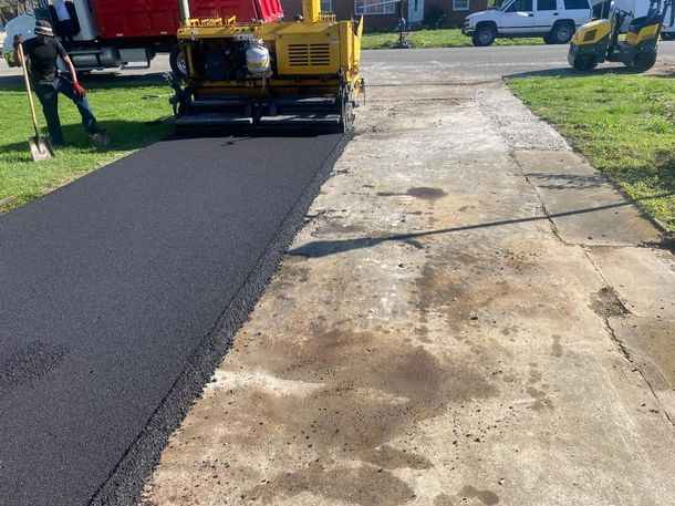 Road paver laying fresh black asphalt beside an old cracked concrete driveway