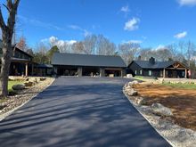 Long black driveway leading to a modern house and garage under a blue sky, with trees and landscaping.