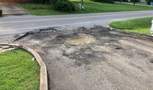Large pothole-ridden asphalt driveway with standing water near a curb and road