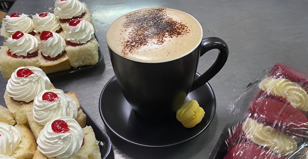 A Cup Of Coffee Is Sitting On A Saucer Next To A Tray Of Desserts — Cedar Park Bakehouse in Taranganba, QLD