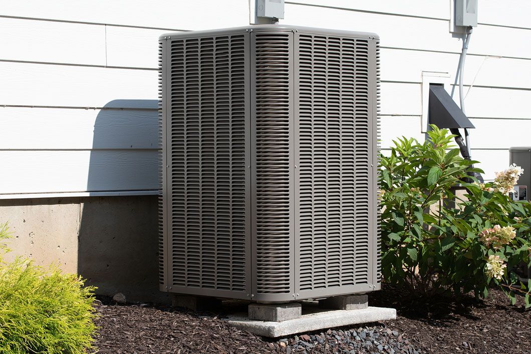 Air conditioning unit outside a white building, on concrete blocks, next to bushes.
