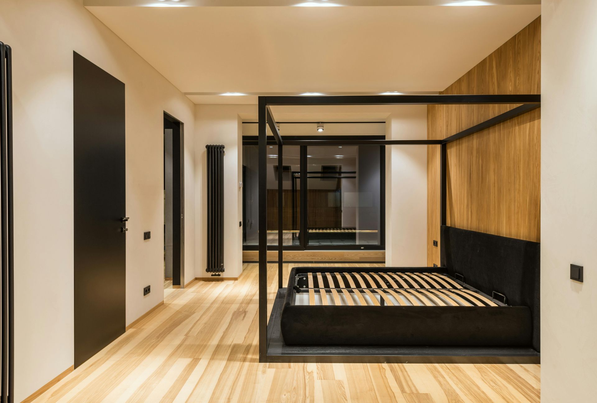 Modern bedroom with four-poster bed, wood floors, wood panel accent wall, and black doors.