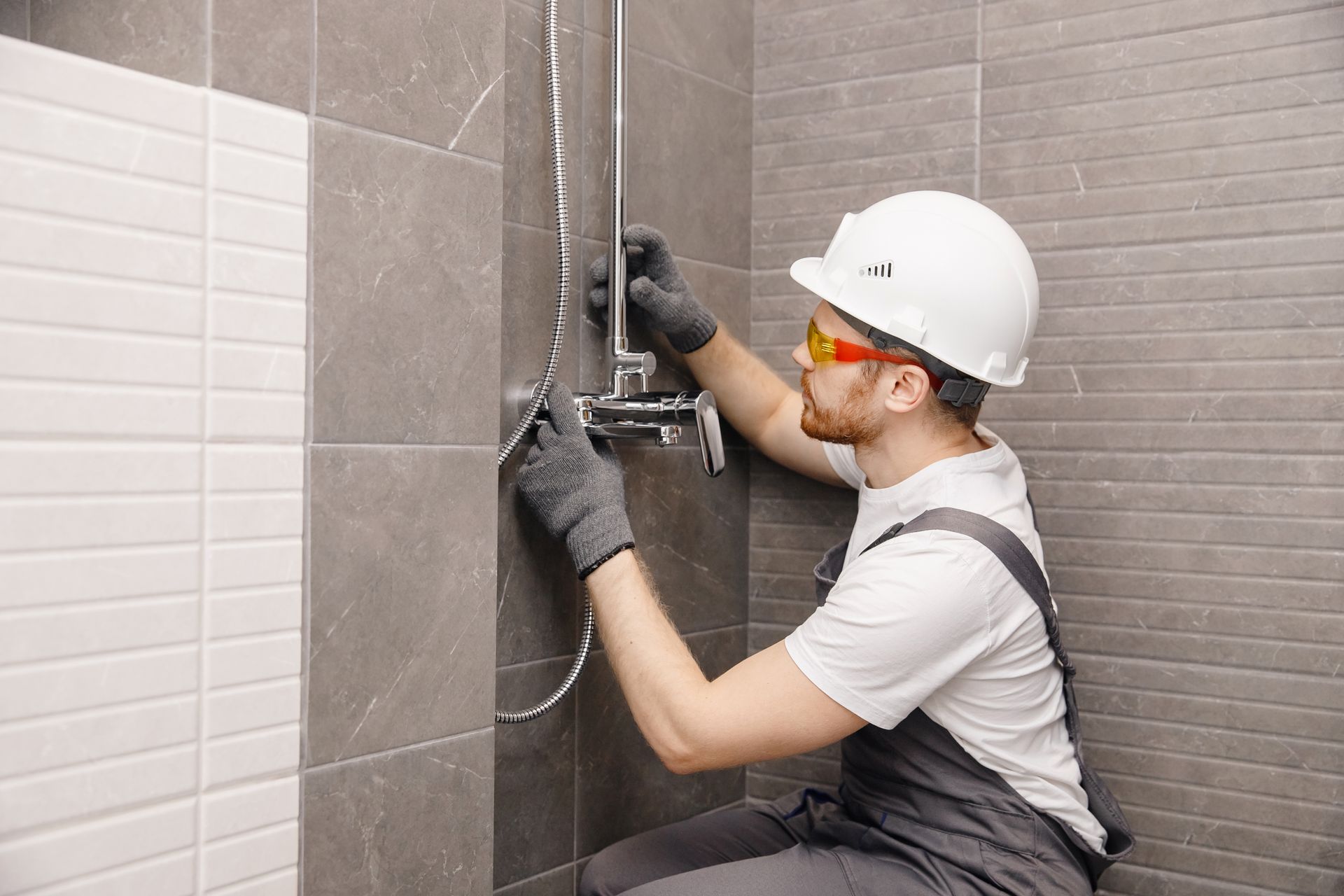 Worker tiling a bathroom wall, wearing a hard hat, gloves, and safety glasses.