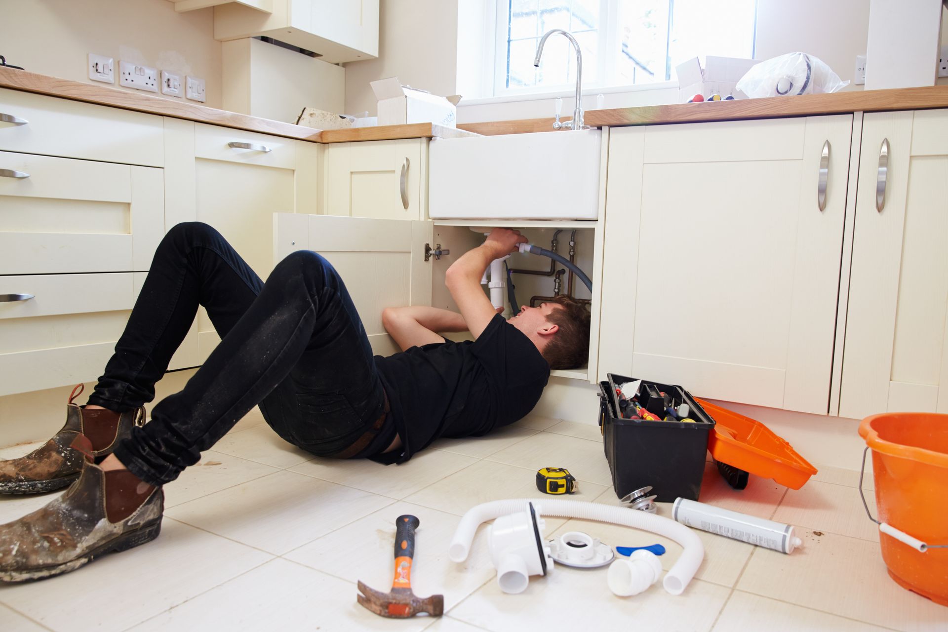 Person lying under a kitchen sink, using tools to fix plumbing, with supplies scattered on the floor. Person lying under a kitchen sink, using tools to fix plumbing, with supplies scattered on the floor.