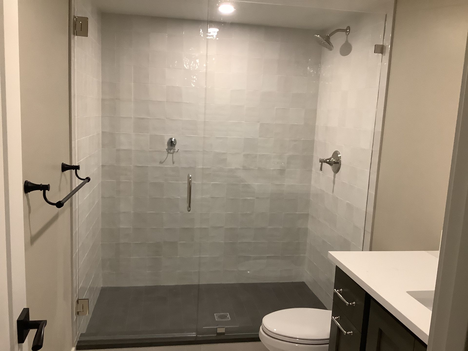 Technician lying under a sink, tightening plumbing with tools beside him in a kitchen. Bathroom with walk-in shower, glass enclosure, and toilet beside a vanity counter.