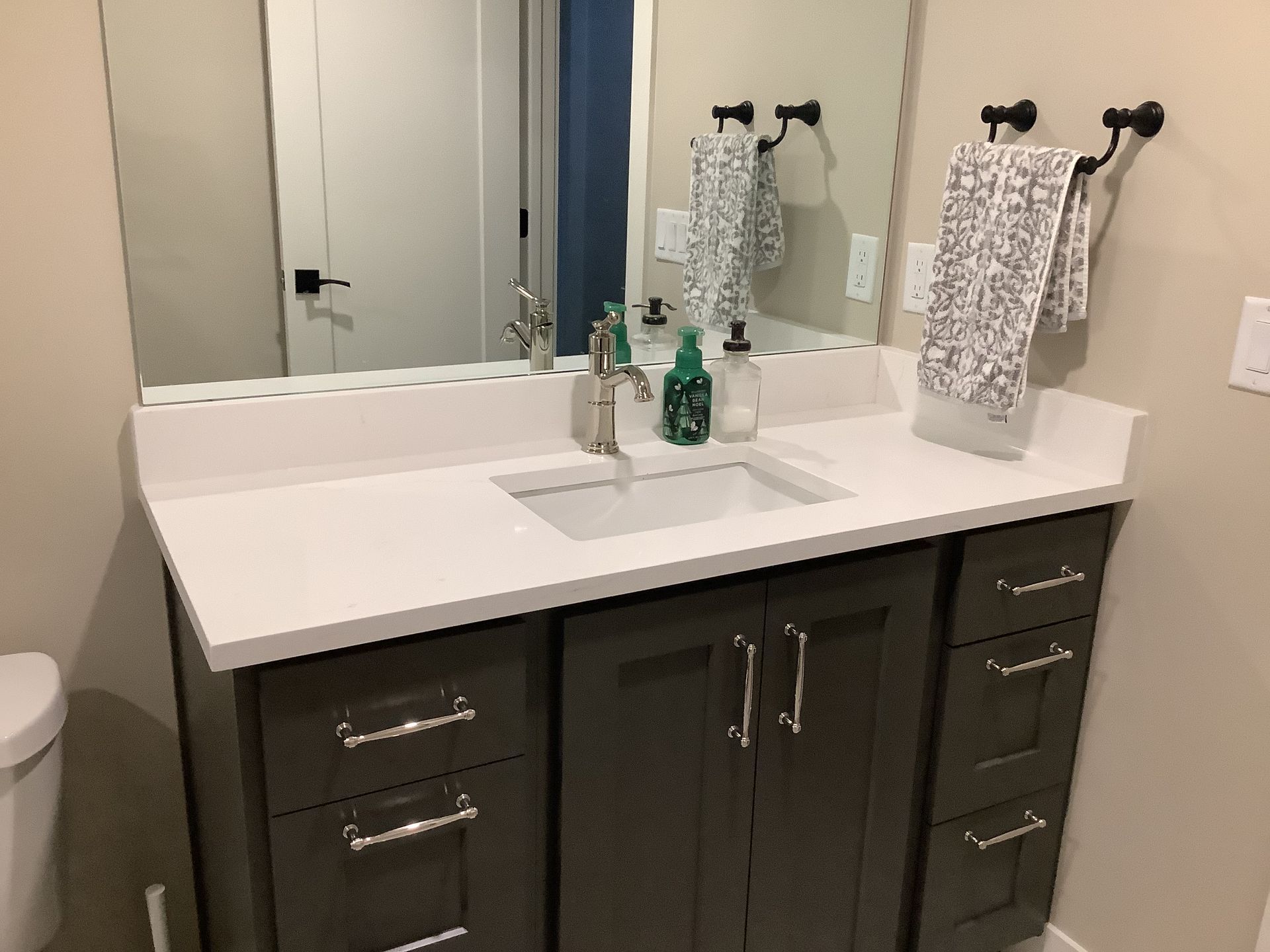 Technician repairing plumbing under a kitchen sink with tools in a white cabinet Bathroom vanity with white countertop, dark cabinets, mirror, and towels hanging on the wall