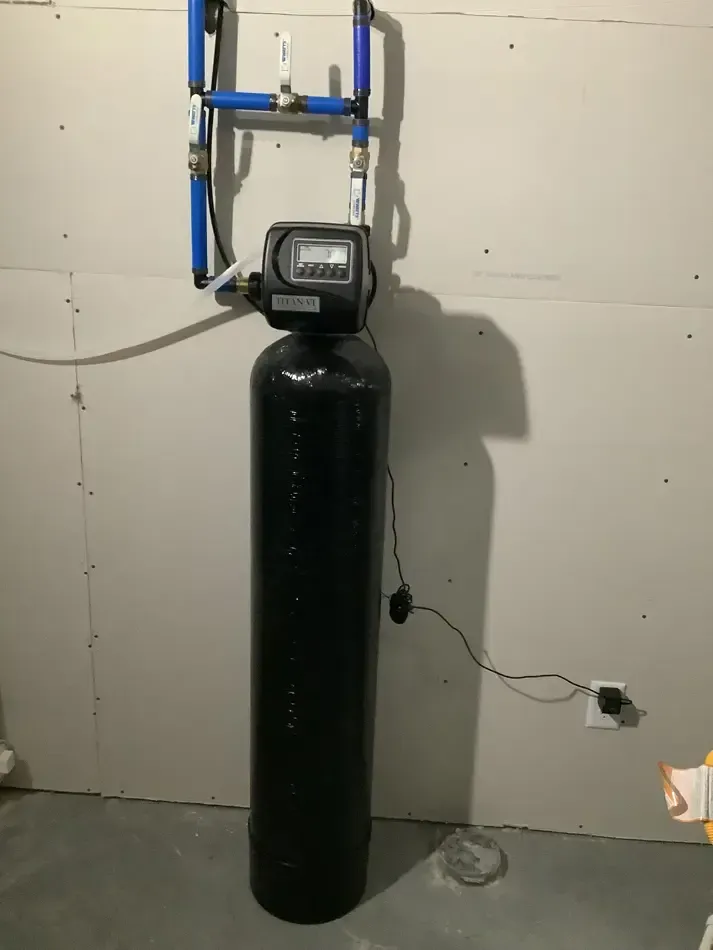 Black water softener tank with control valve in a utility room, mounted pipes above and a power cord beside it