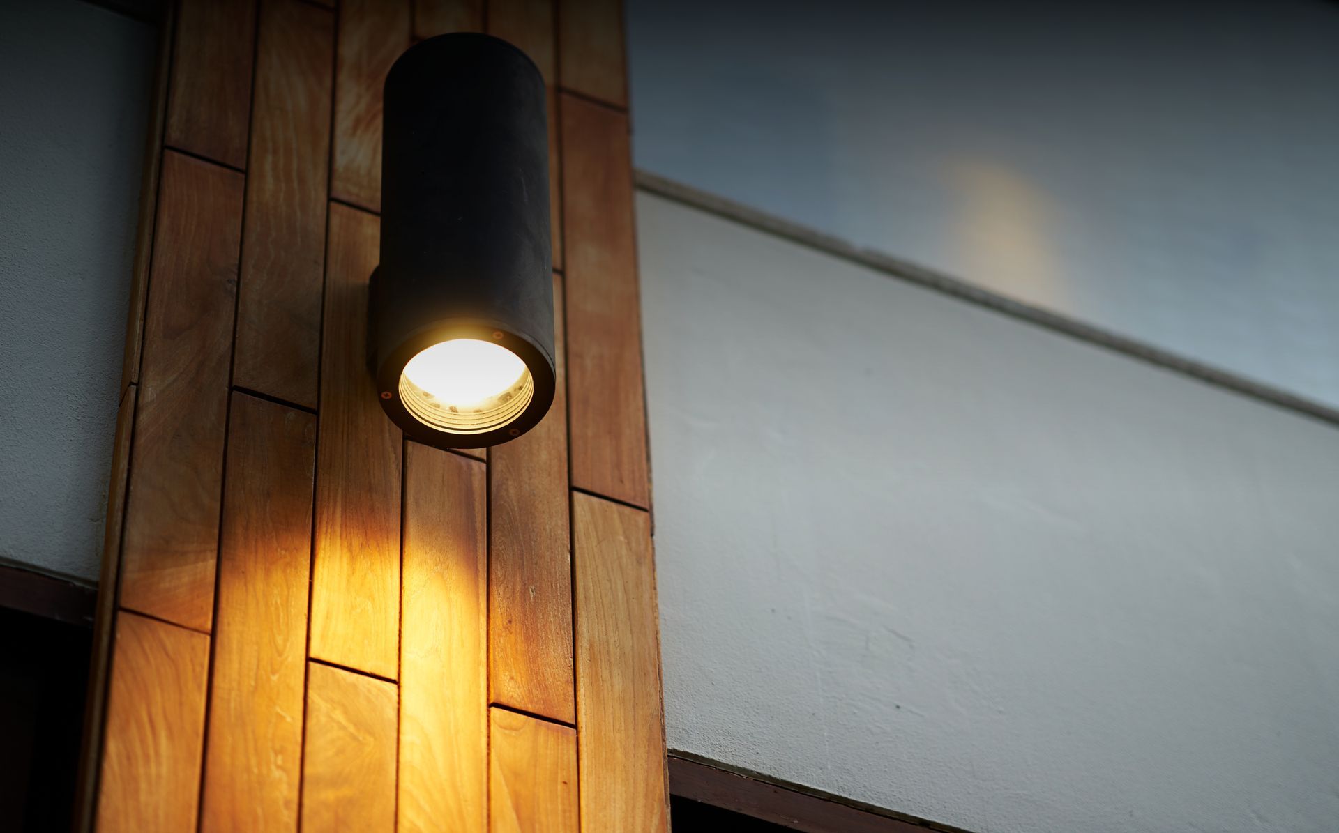 Black cylinder light fixture on wooden wall, casting light on the wood.