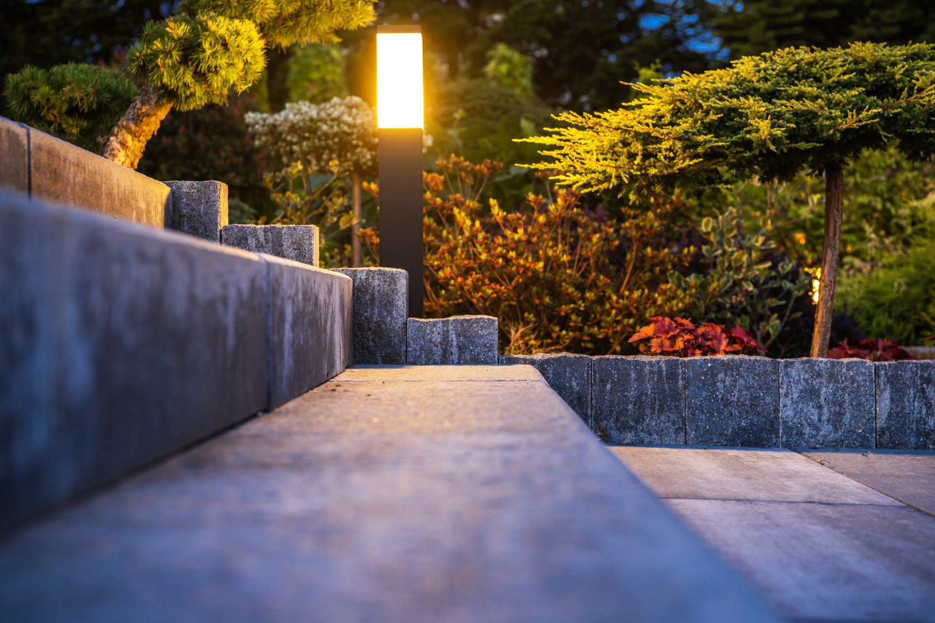 Stone steps lead up to a lit outdoor lamp. Lush greenery surrounds the steps and lamp.