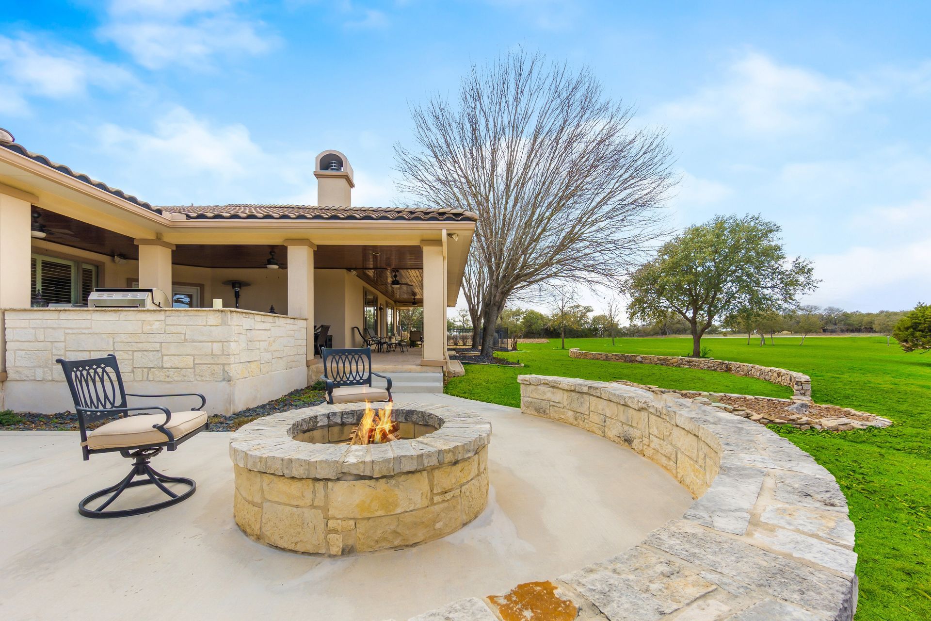 Patio with fire pit, stone seating, and a view of a green field under a blue sky.