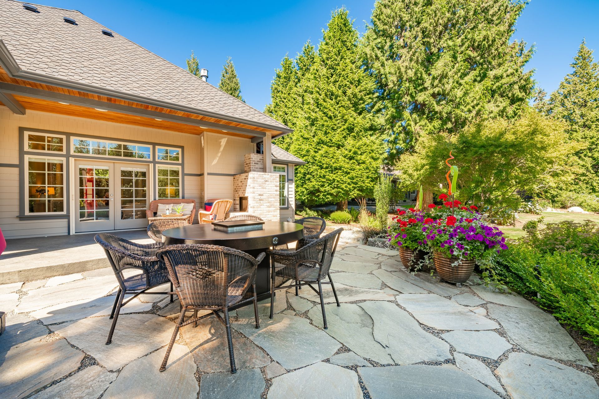 Patio with round table and chairs, flowers, and a house with glass doors, set in a sunny garden.