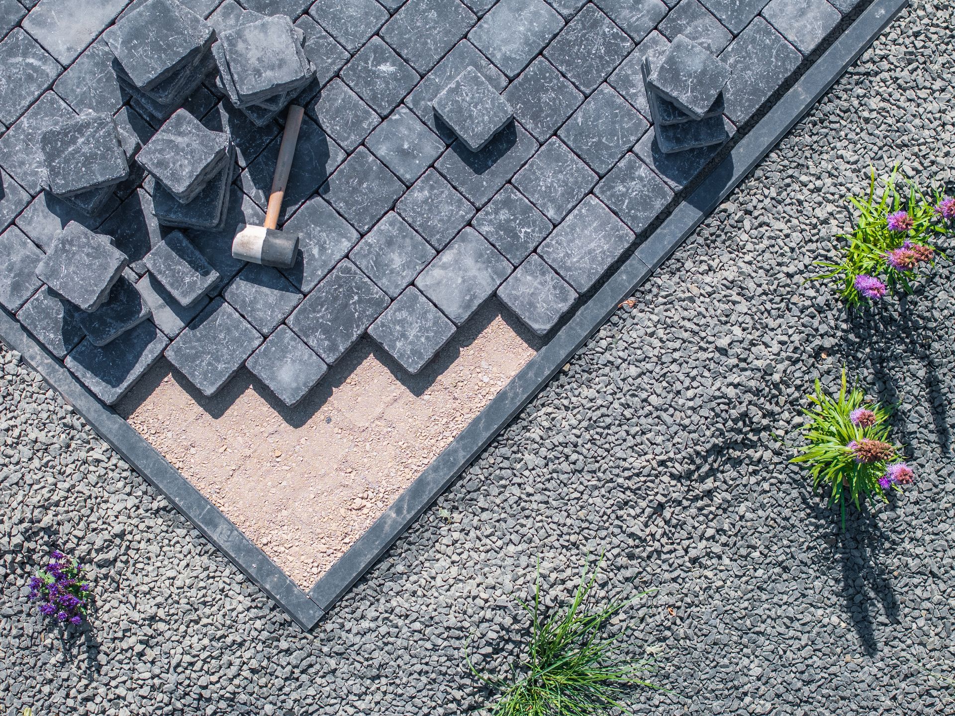 Stone pavers being installed on a gravel base, with a mallet and piles of stones.